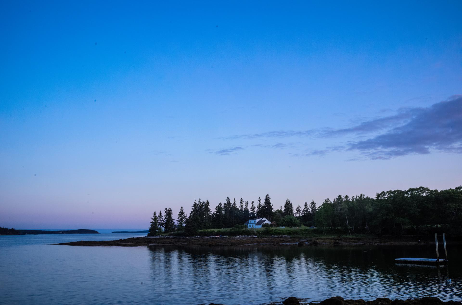 Dusk scene of a calm bay, forested point with a house, under a blue sky.