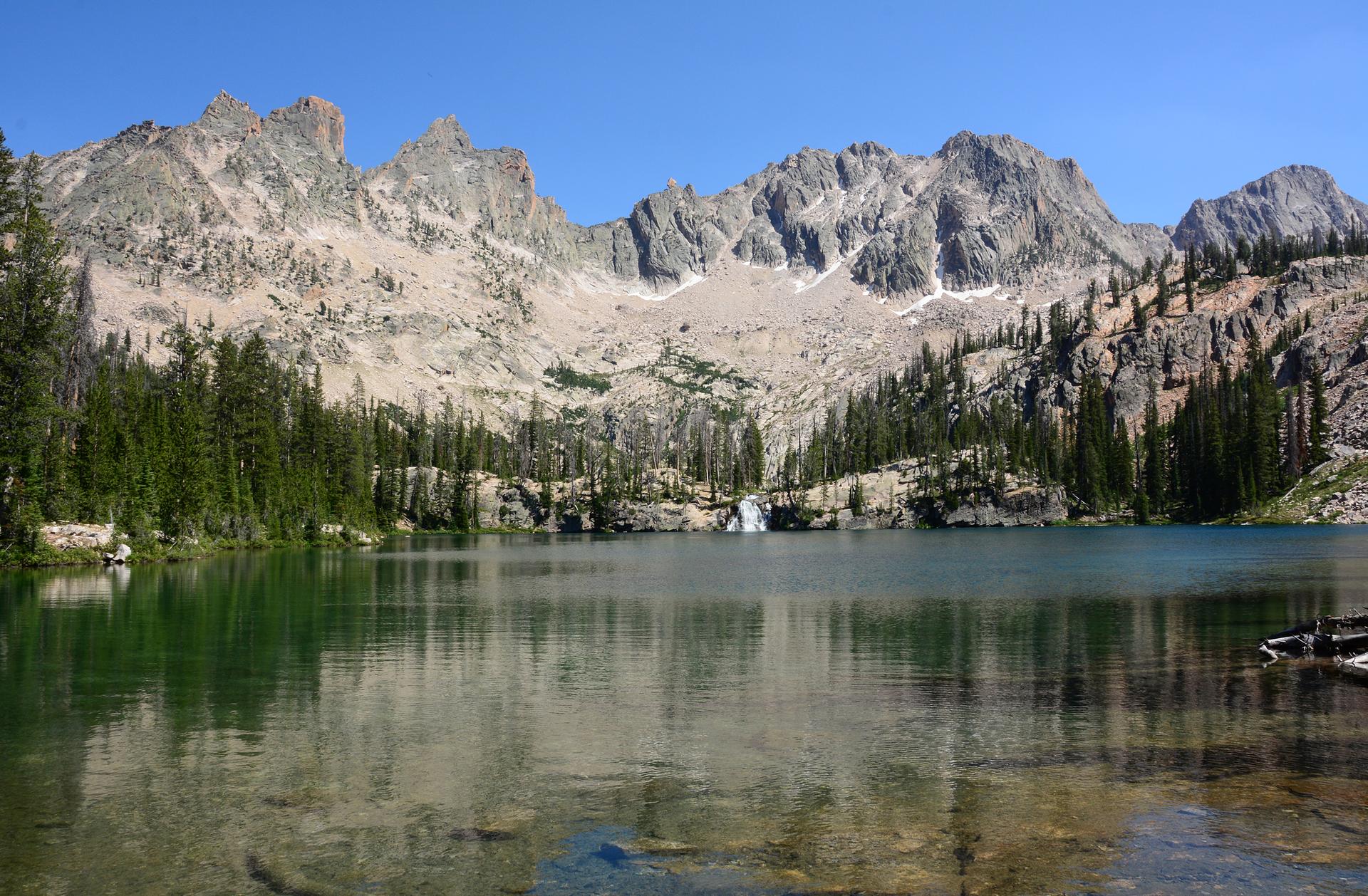 Calm mountain lake reflecting rugged peaks and pine forests under clear blue sky.