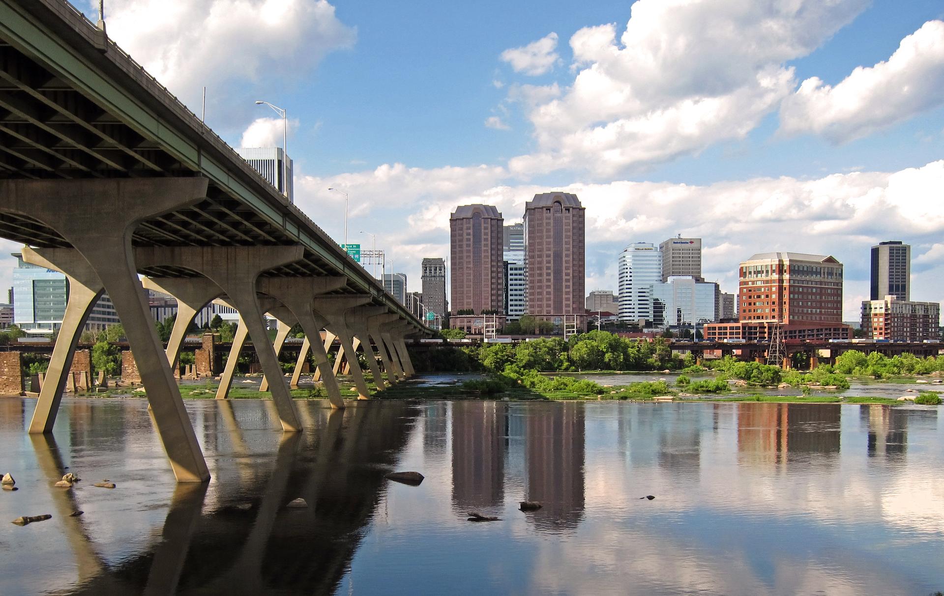 Concrete bridge over a river reflecting a city skyline under a sunny sky.