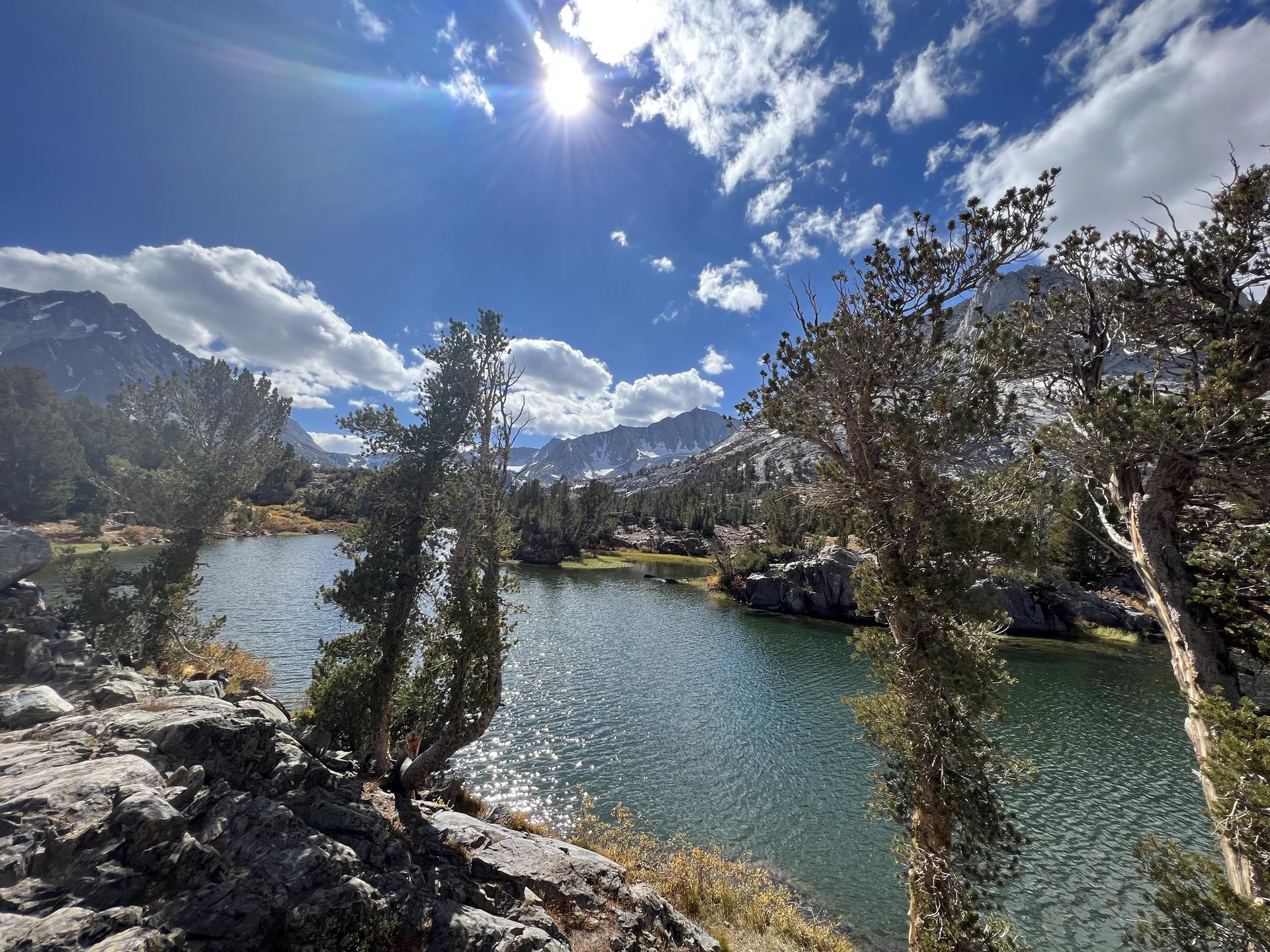 Sunny day over a mountain lake framed by pine trees and clouds.