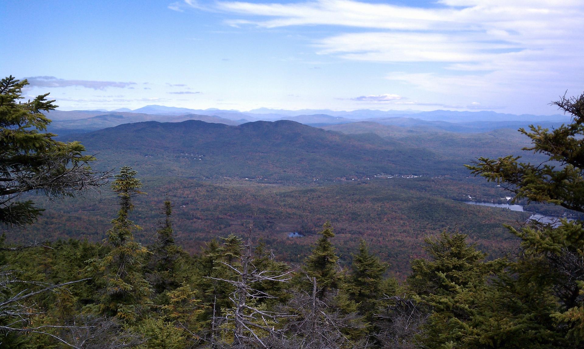 Expansive view of forested mountains under a bright sky, with distant water.