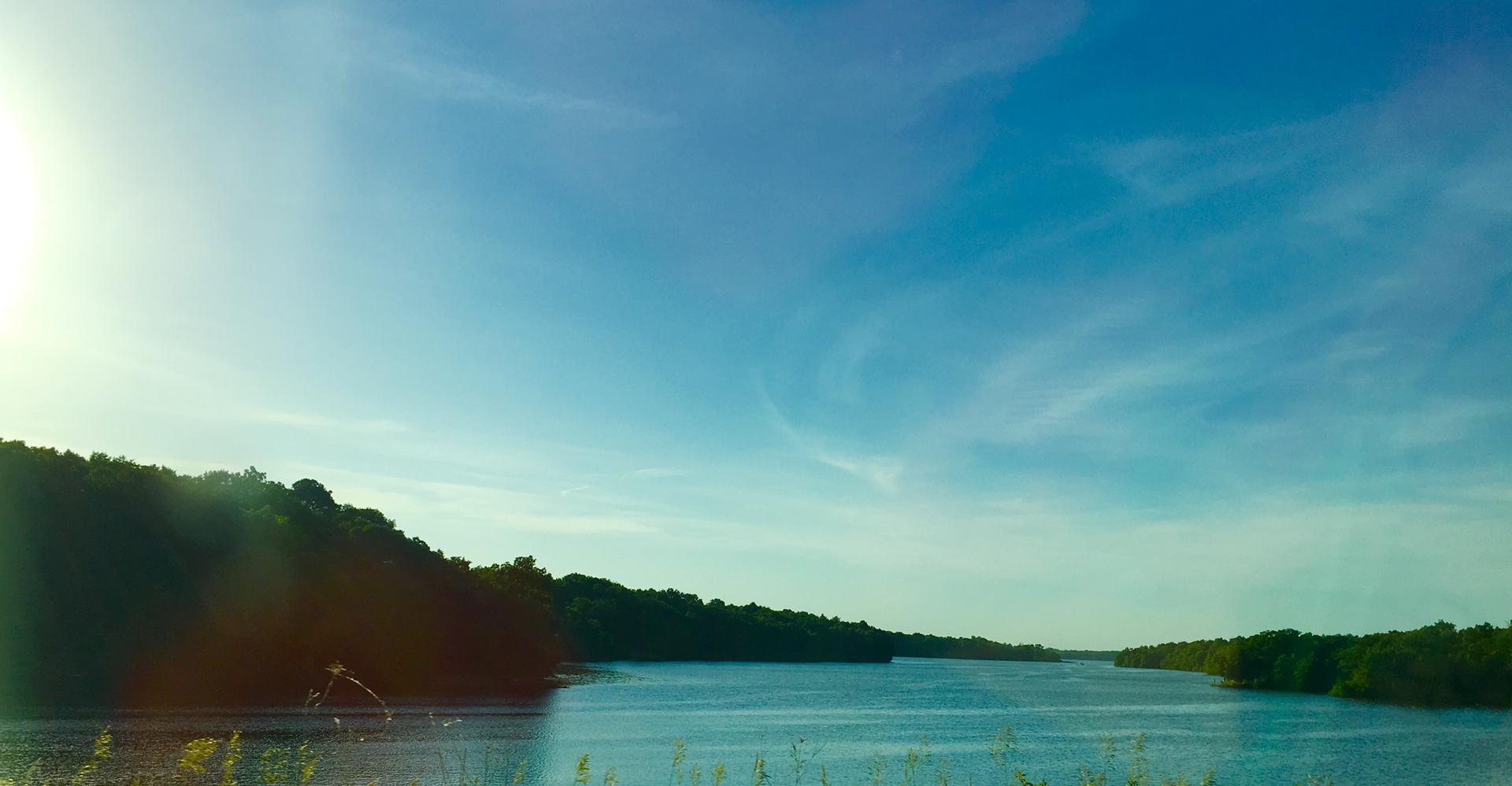 Bright sun over a wide river flanked by green, forested hills.