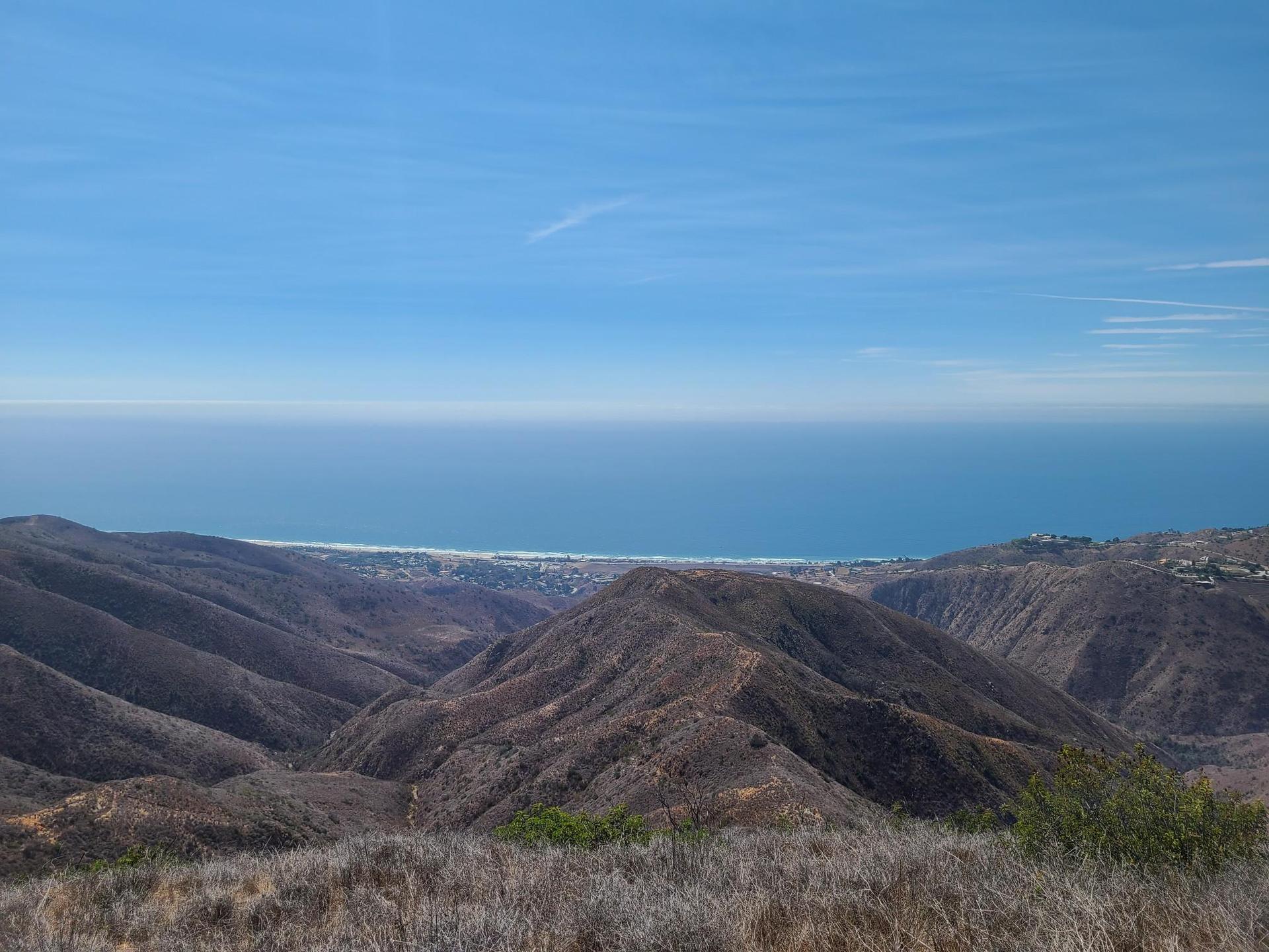 Panoramic view of dry mountains descending to a distant ocean under a clear sky.