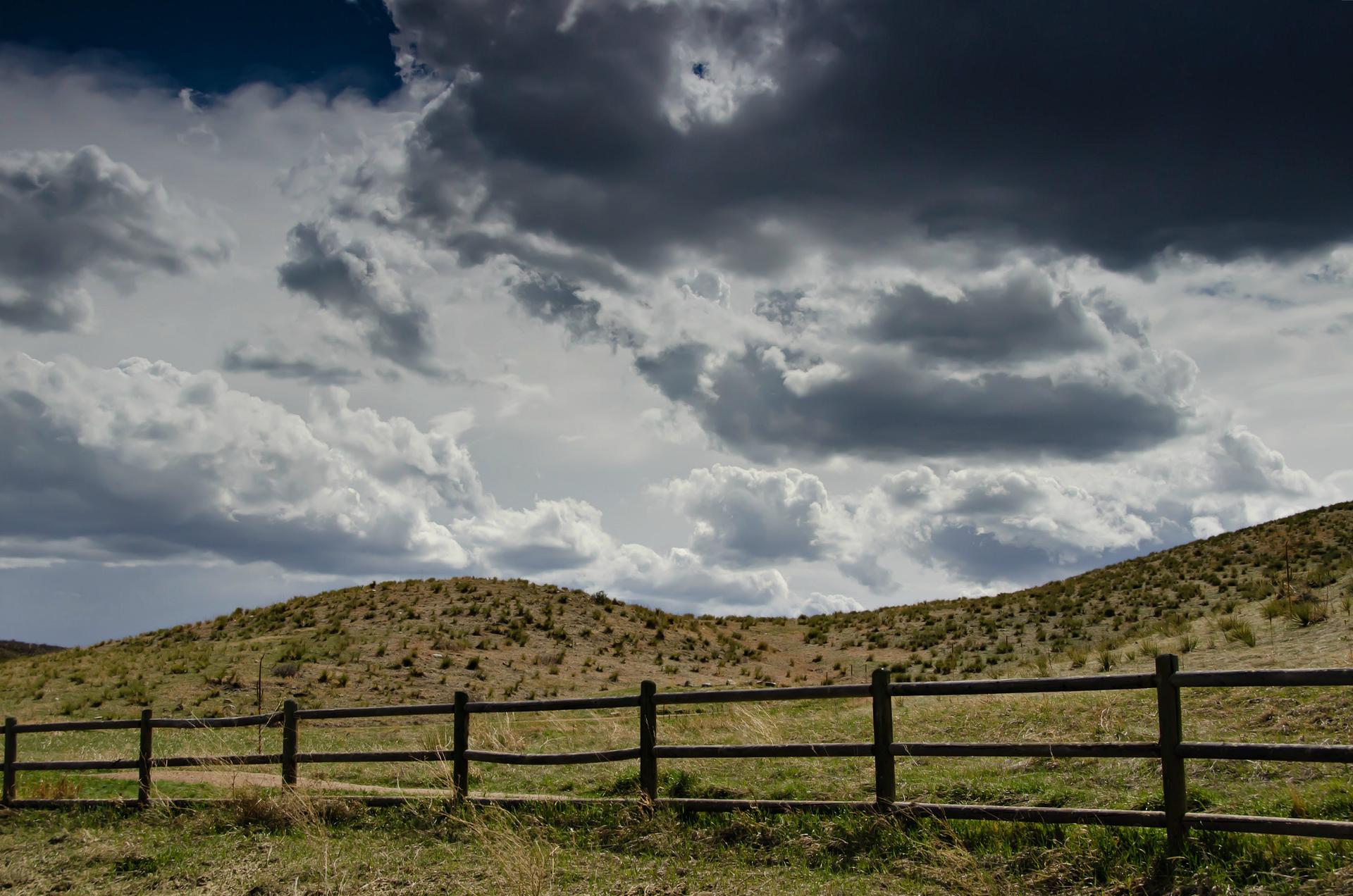 A wooden fence stretches across dry, rolling hills under a dramatic cloudy sky.