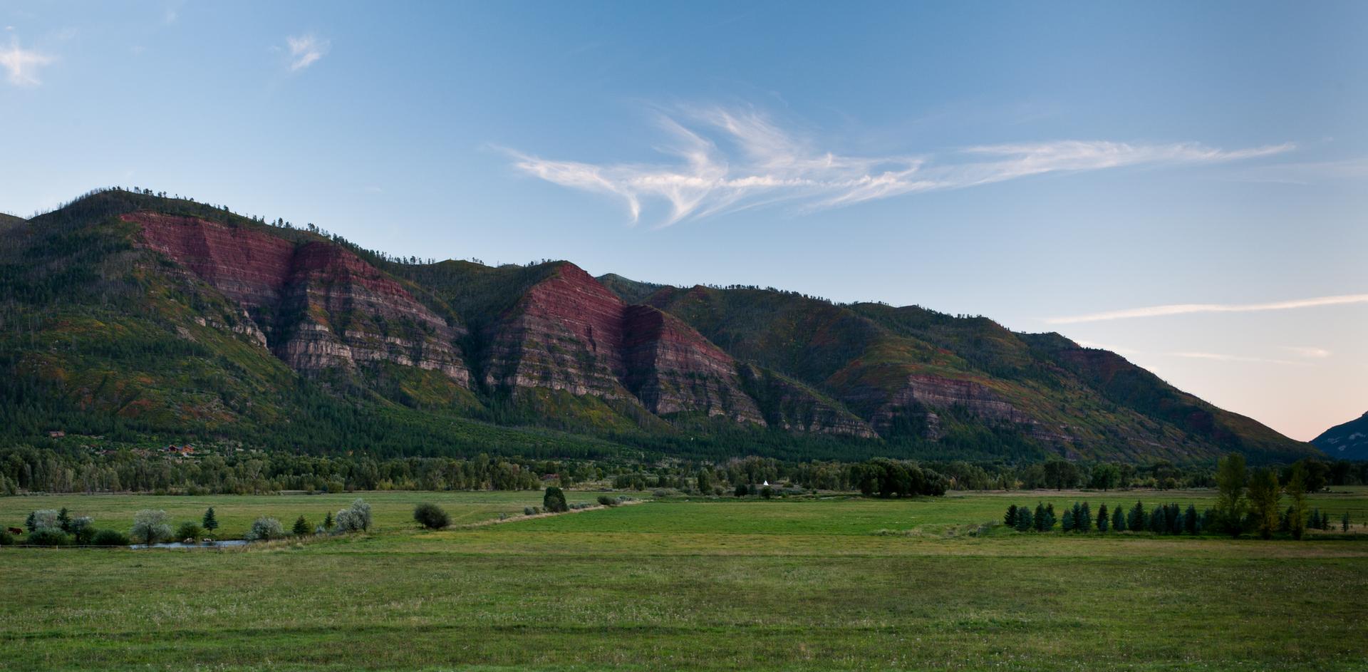 Expansive green fields before layered mountains under a clear blue sky.