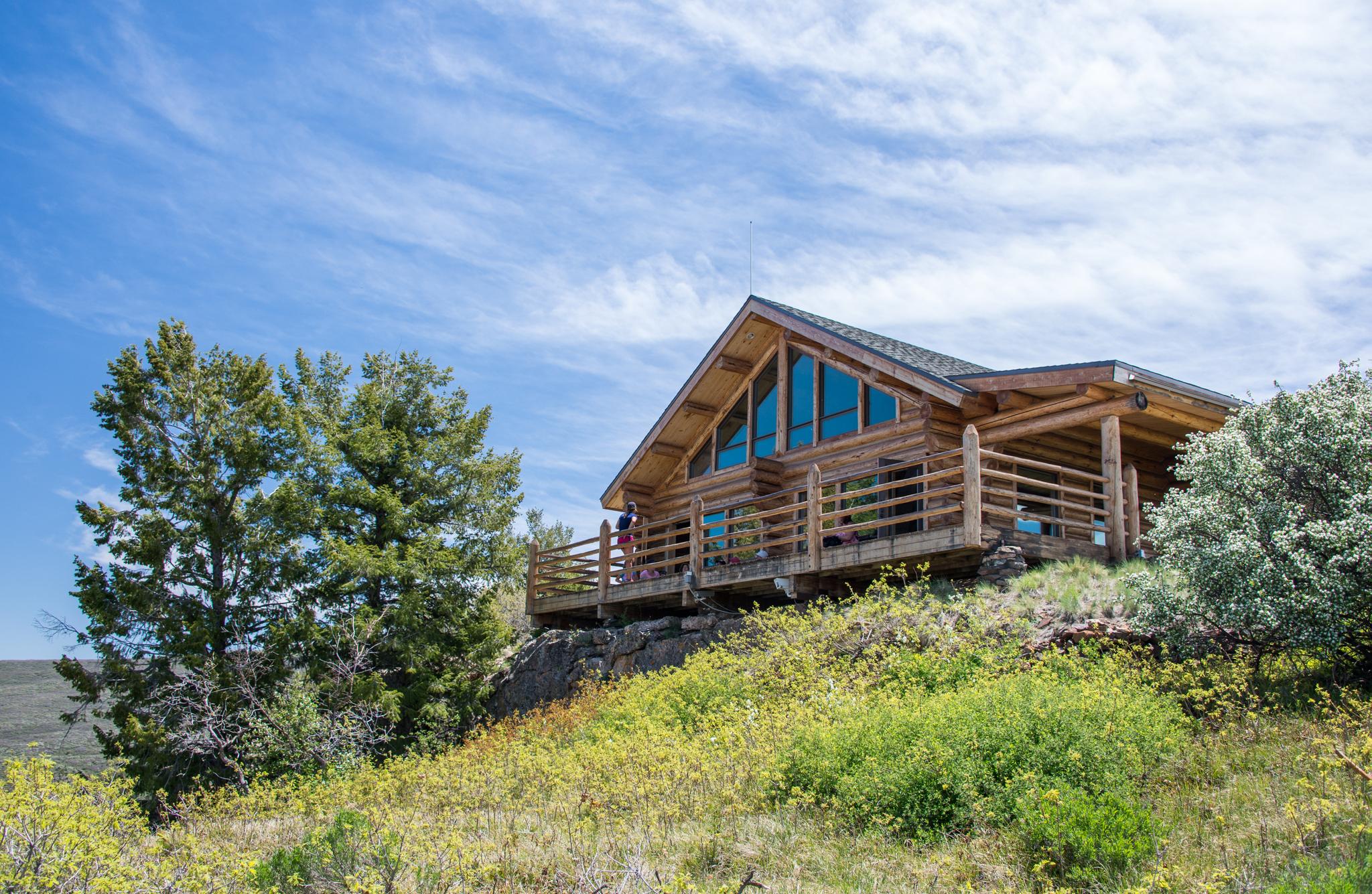 Rustic log cabin with a deck on a sunny, green hillside under blue sky.
