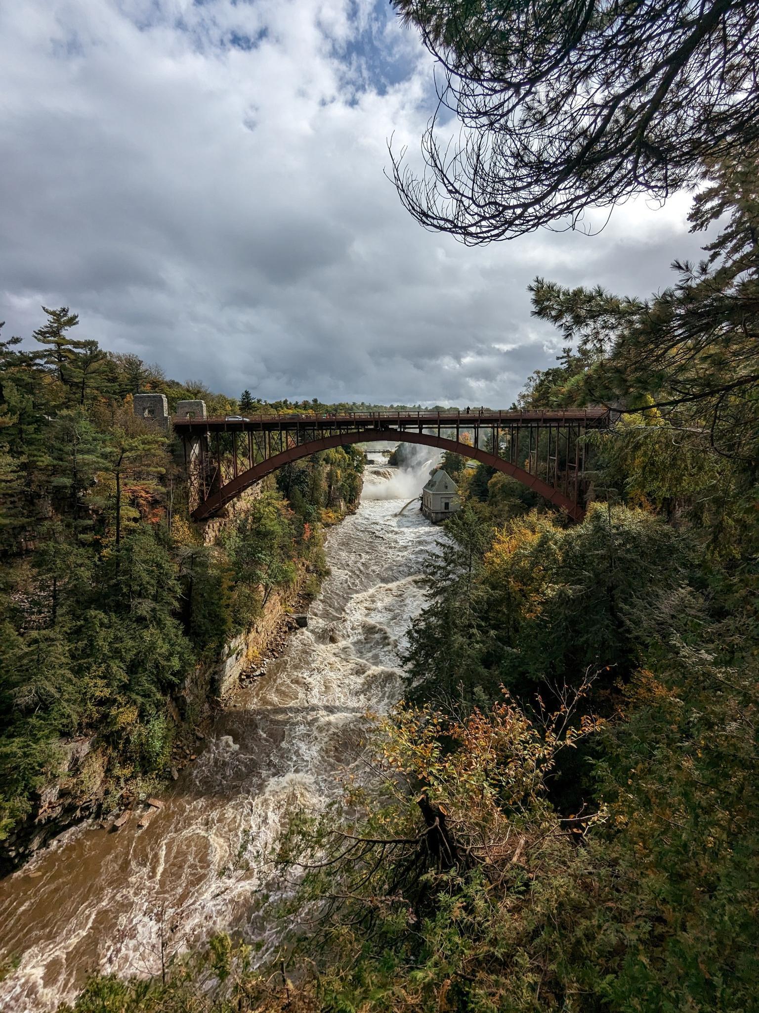 Rushing river flowing under a large metal bridge, surrounded by trees.