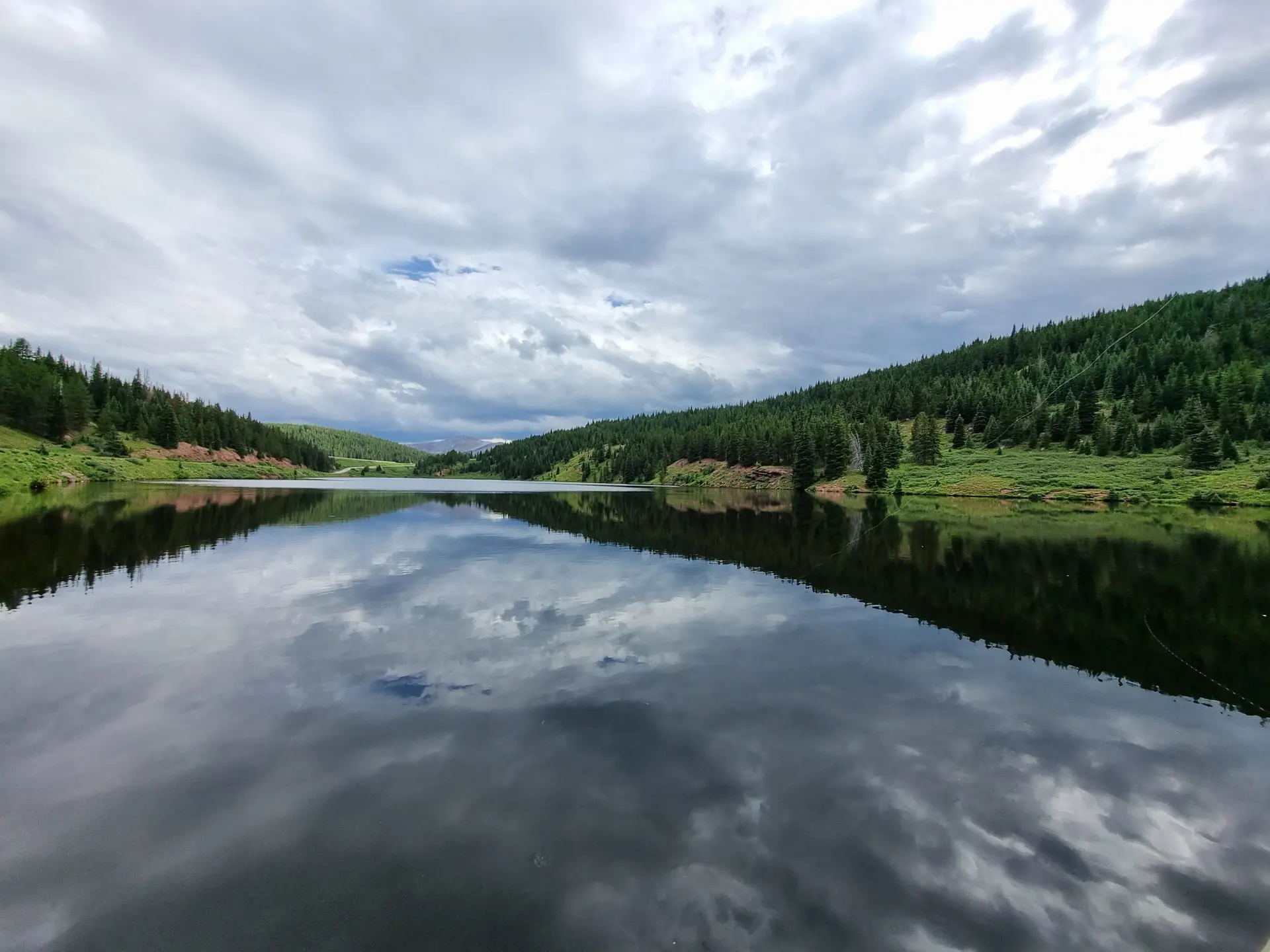 Calm lake reflecting cloudy sky and forested hills, under overcast light.