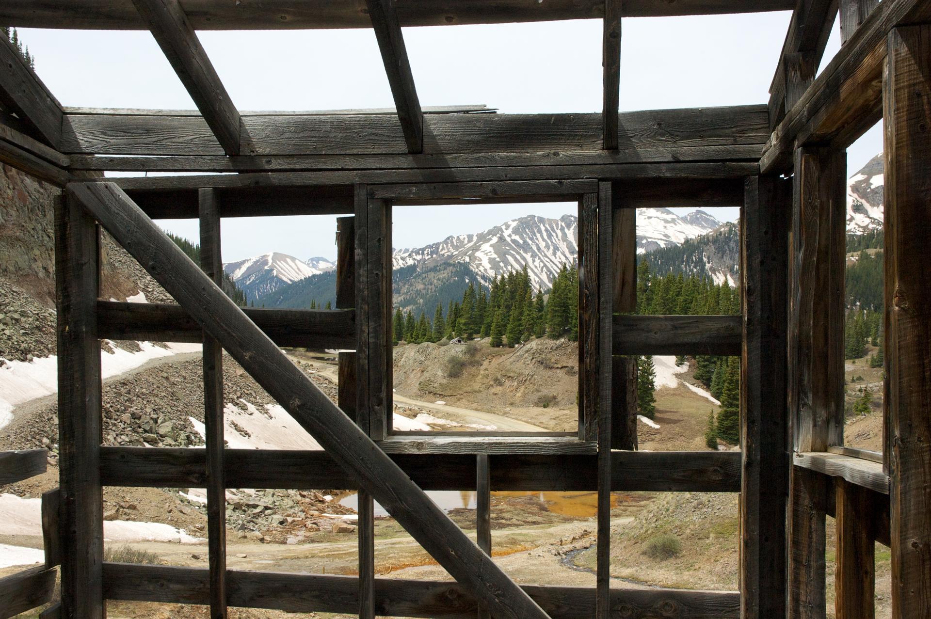 View through old wooden structure to snow-capped mountains and pine trees.