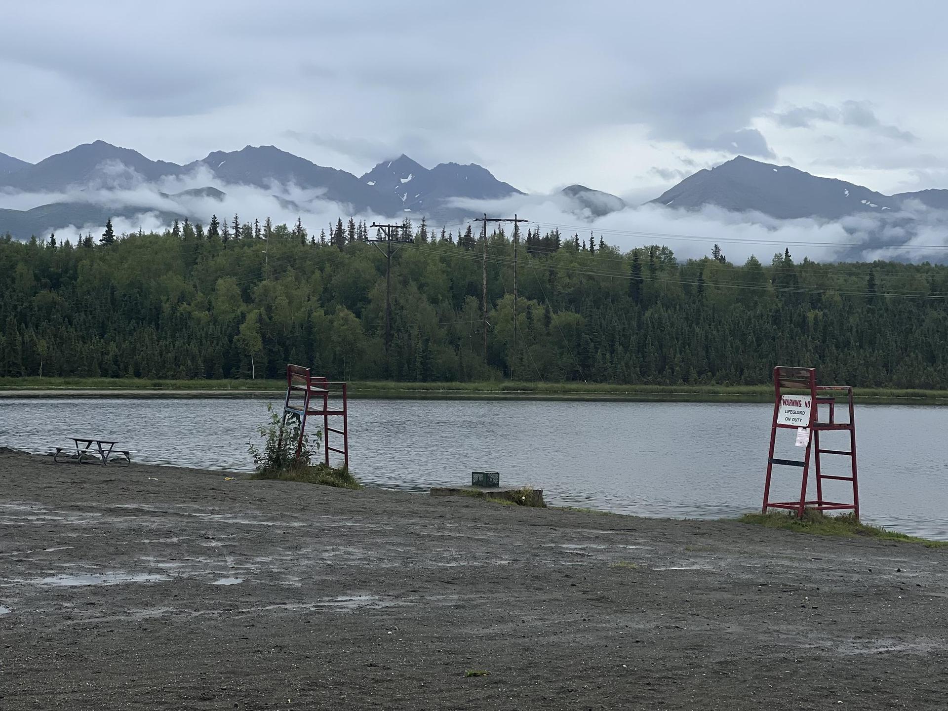 Overcast lake shore with two red lifeguard chairs and cloudy mountains.