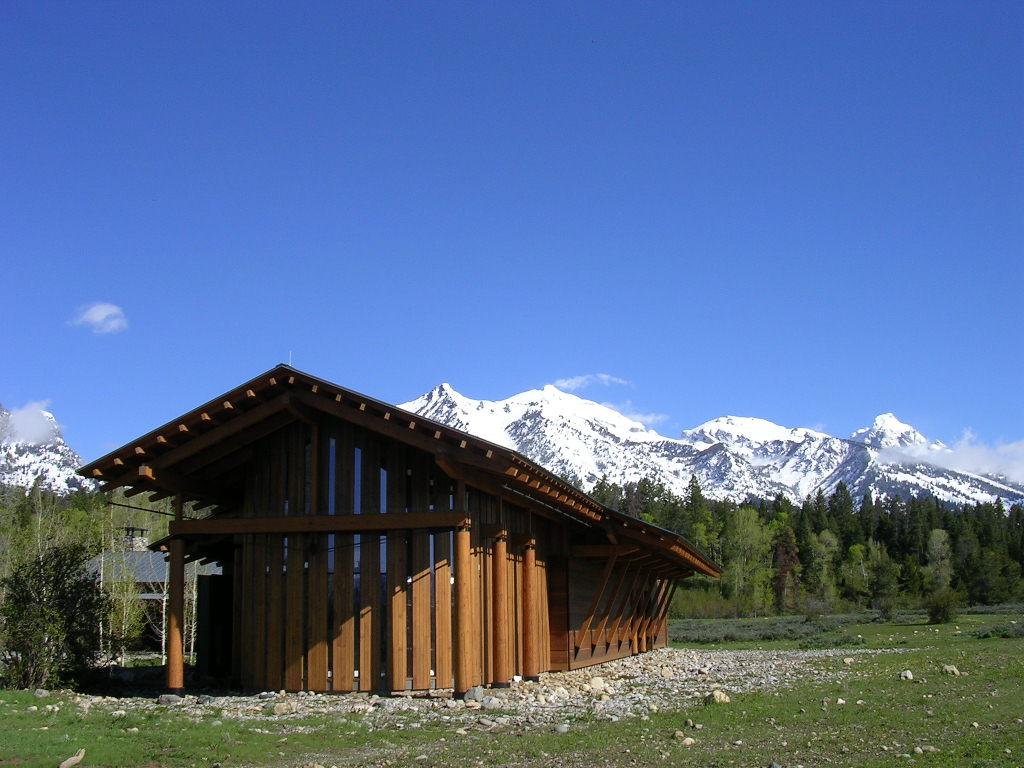 A rustic wooden building in a field with snow-capped mountains behind it.