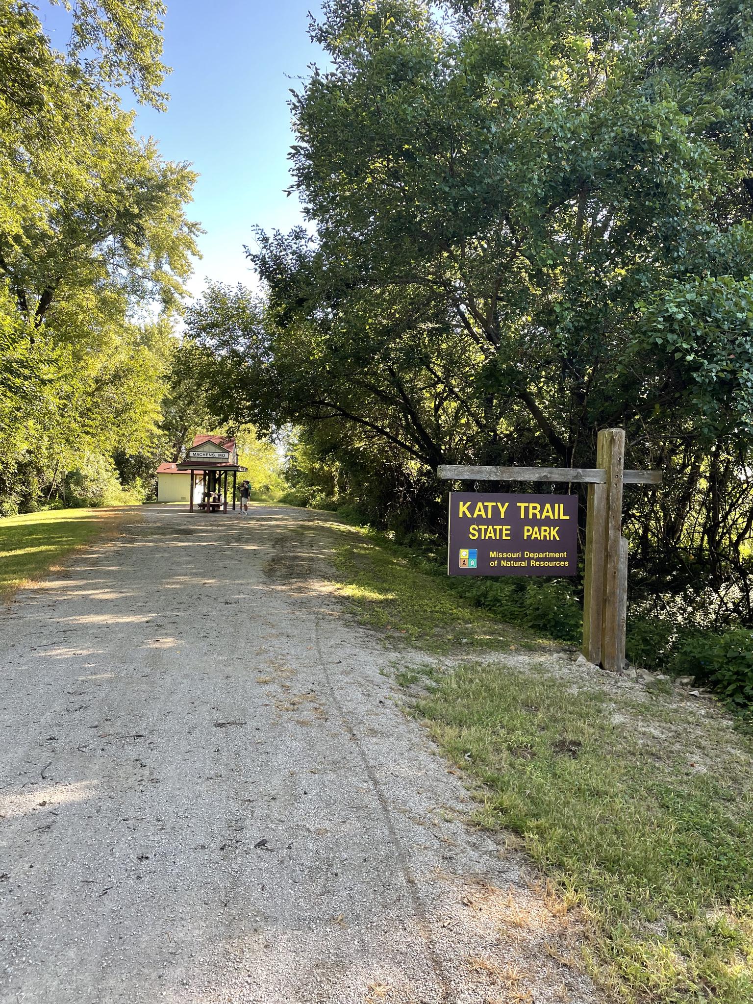 Sunny dirt path leading to a small building, bordered by green trees and a sign.