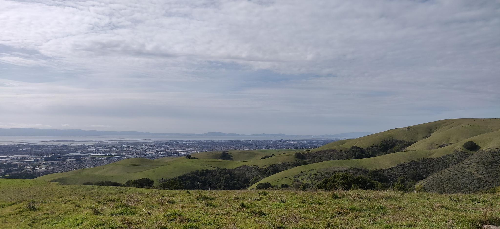 Expansive view of rolling green hills, a distant city, and water under cloudy skies.