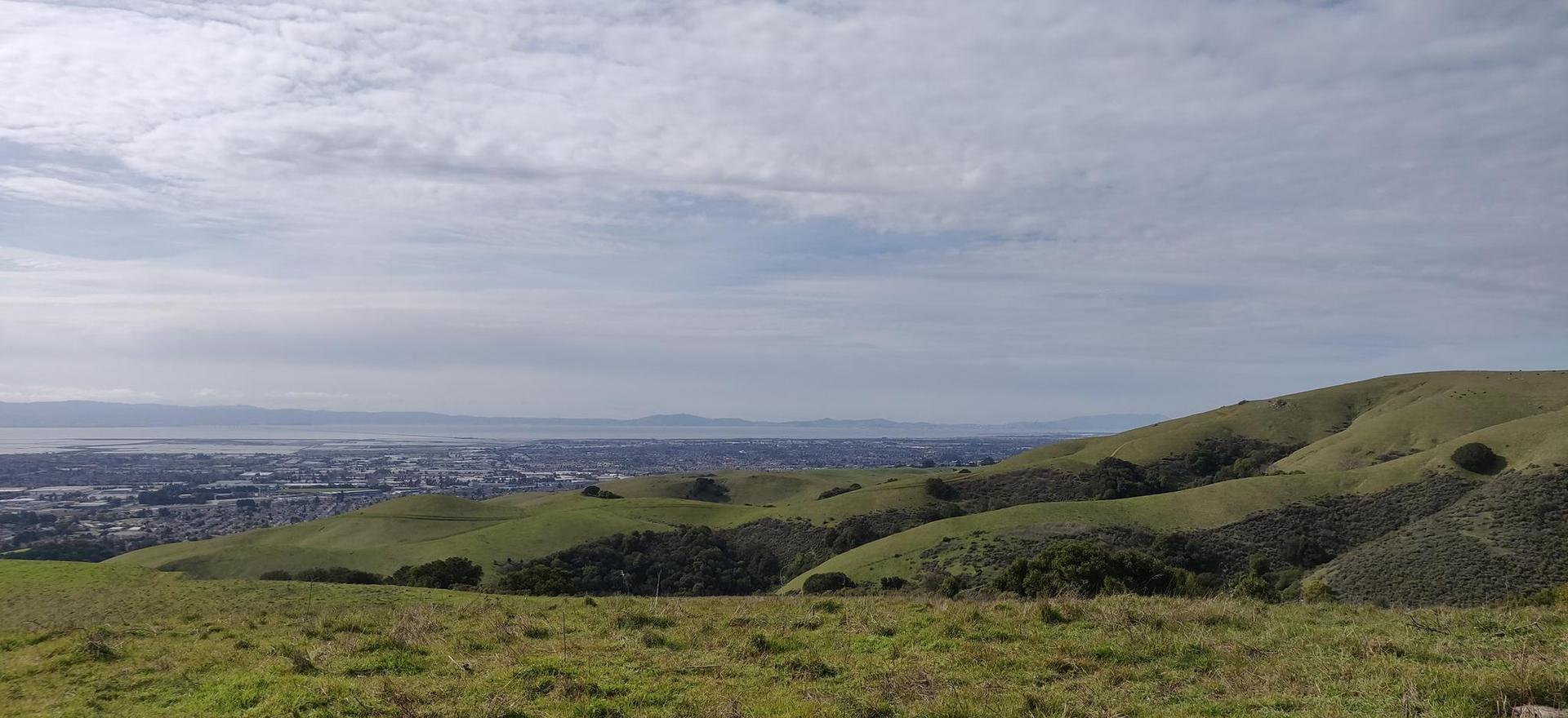 Expansive view of rolling green hills, a distant city, and water under cloudy skies.