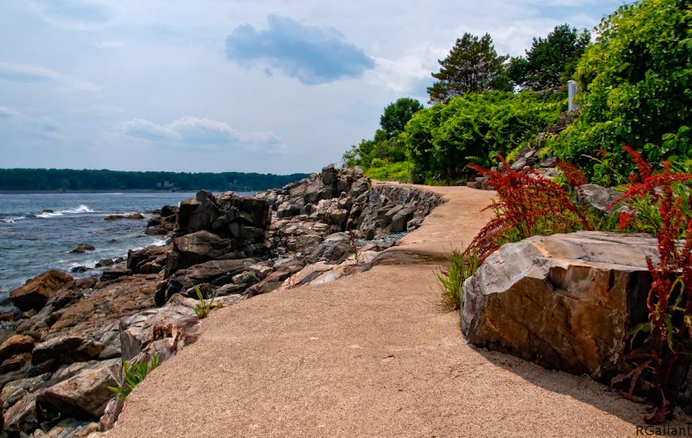 Paved path winding along a rocky ocean coastline under a cloudy sky.