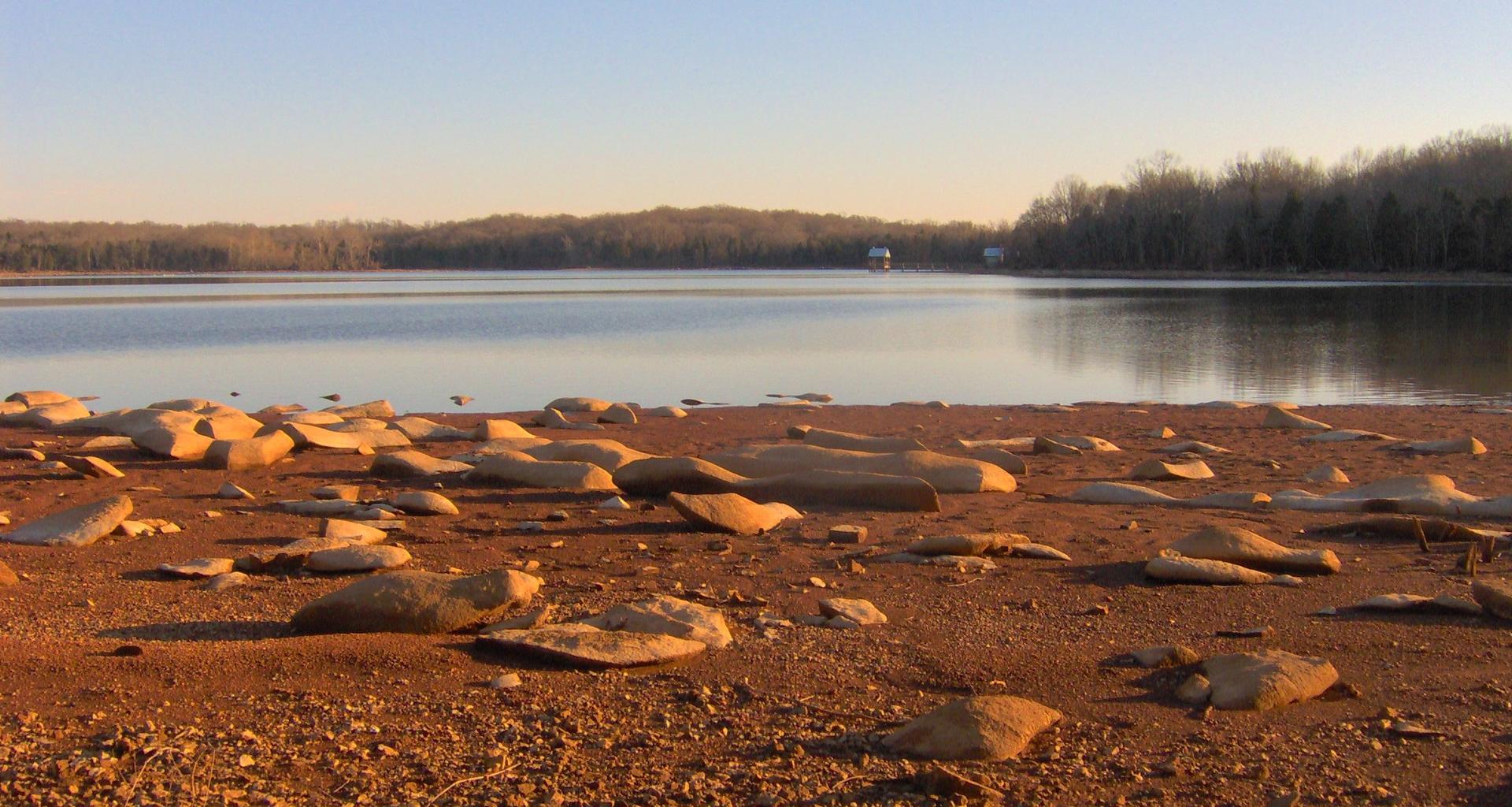 Golden light illuminates rocky lake shore with calm water and distant trees.
