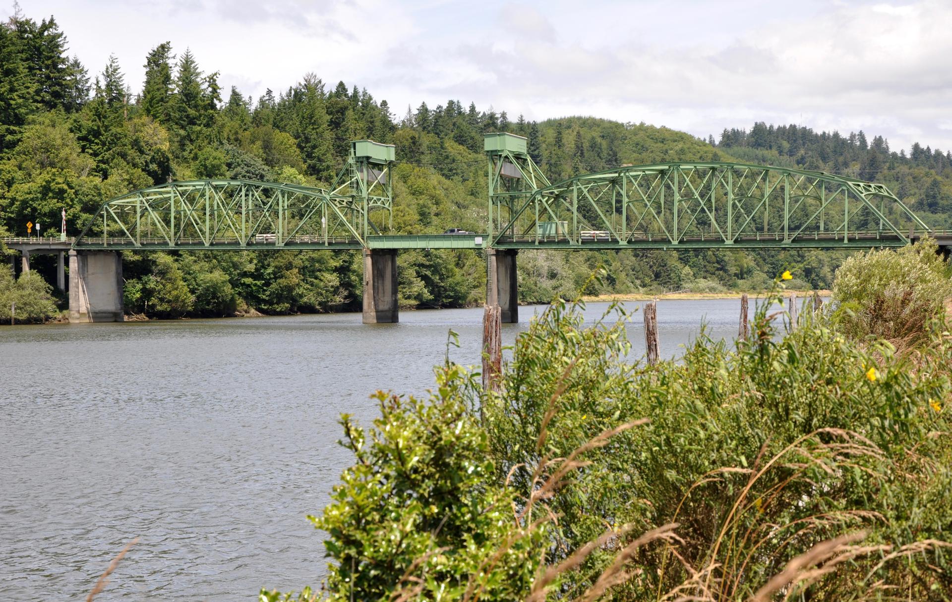 Green truss bridge spanning a river with lush forested hills in background.