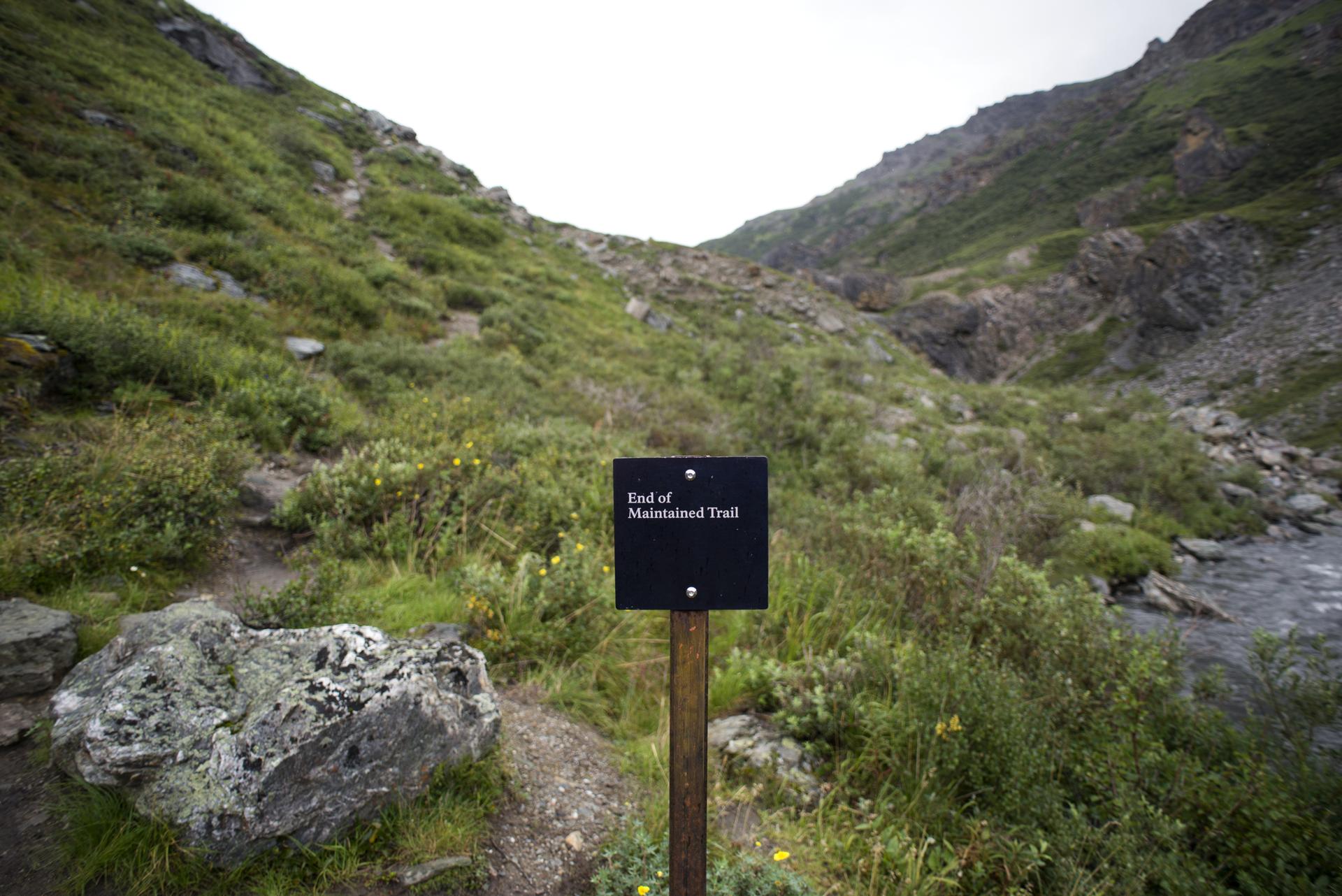 A sign marks the end of a maintained trail in a rocky, green mountain valley.