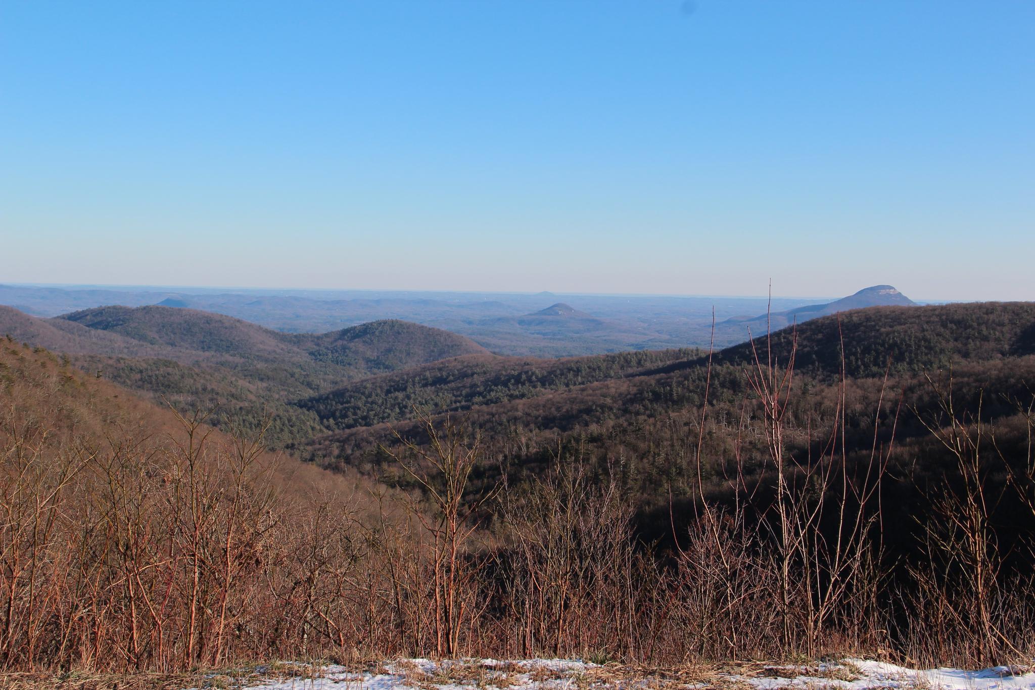 Vast mountain landscape with bare winter trees, distant hazy peaks, and clear sky.