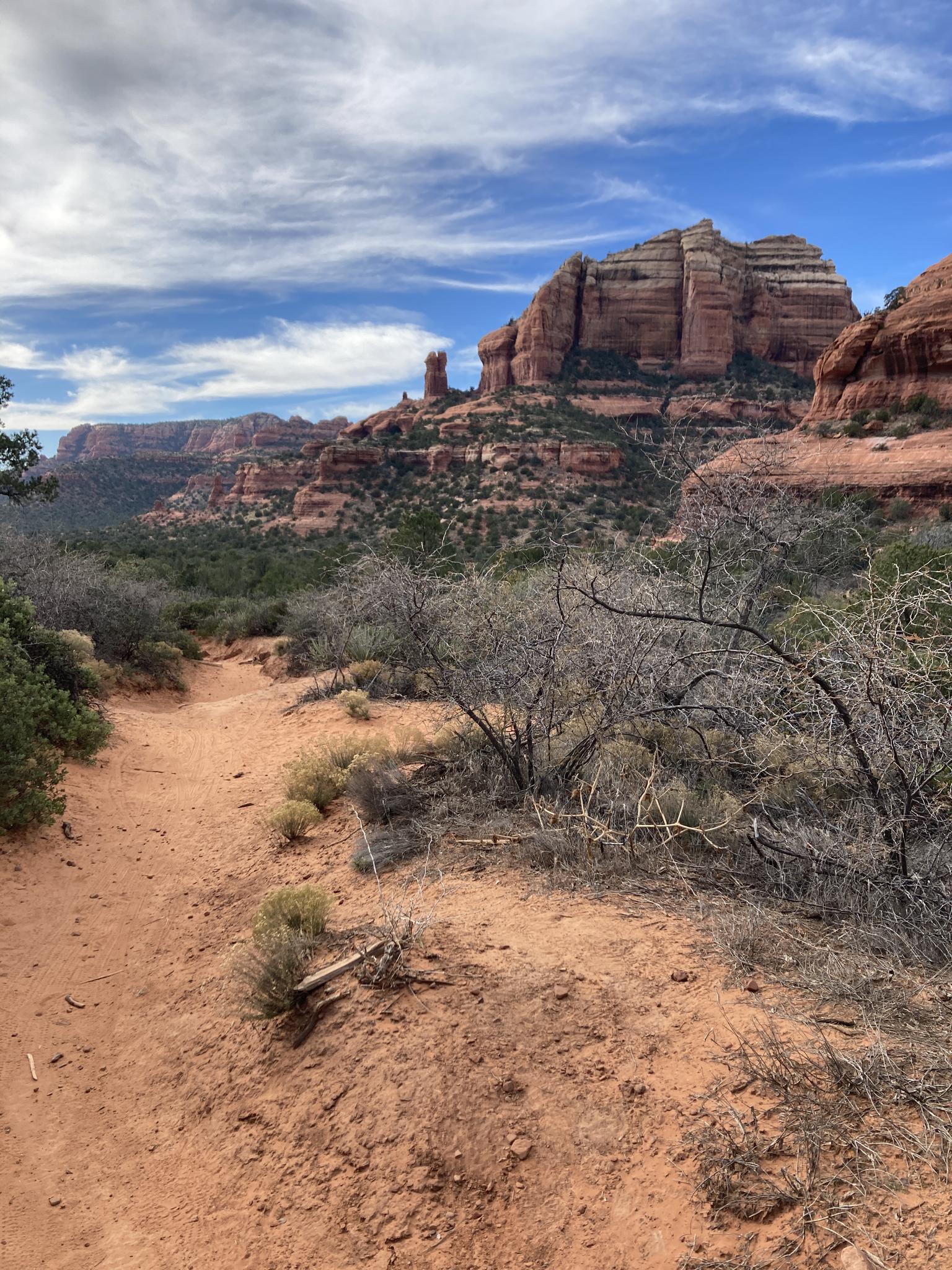 A red dirt path winds through dry brush towards towering red rock formations.