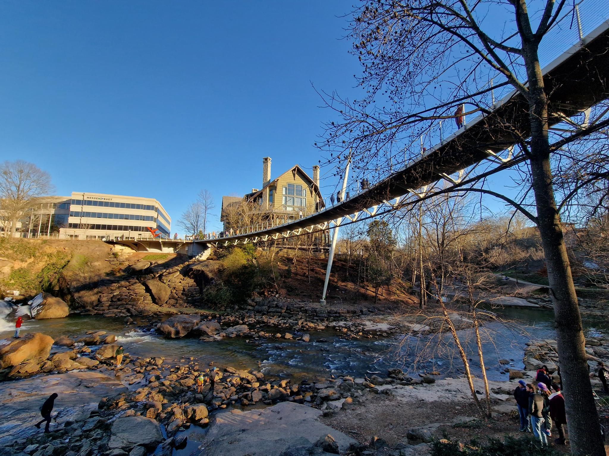 Suspension bridge spans a rocky river with buildings and people under a clear sky. - Paris Mountain and Swamp Rabbit Trail Loop
