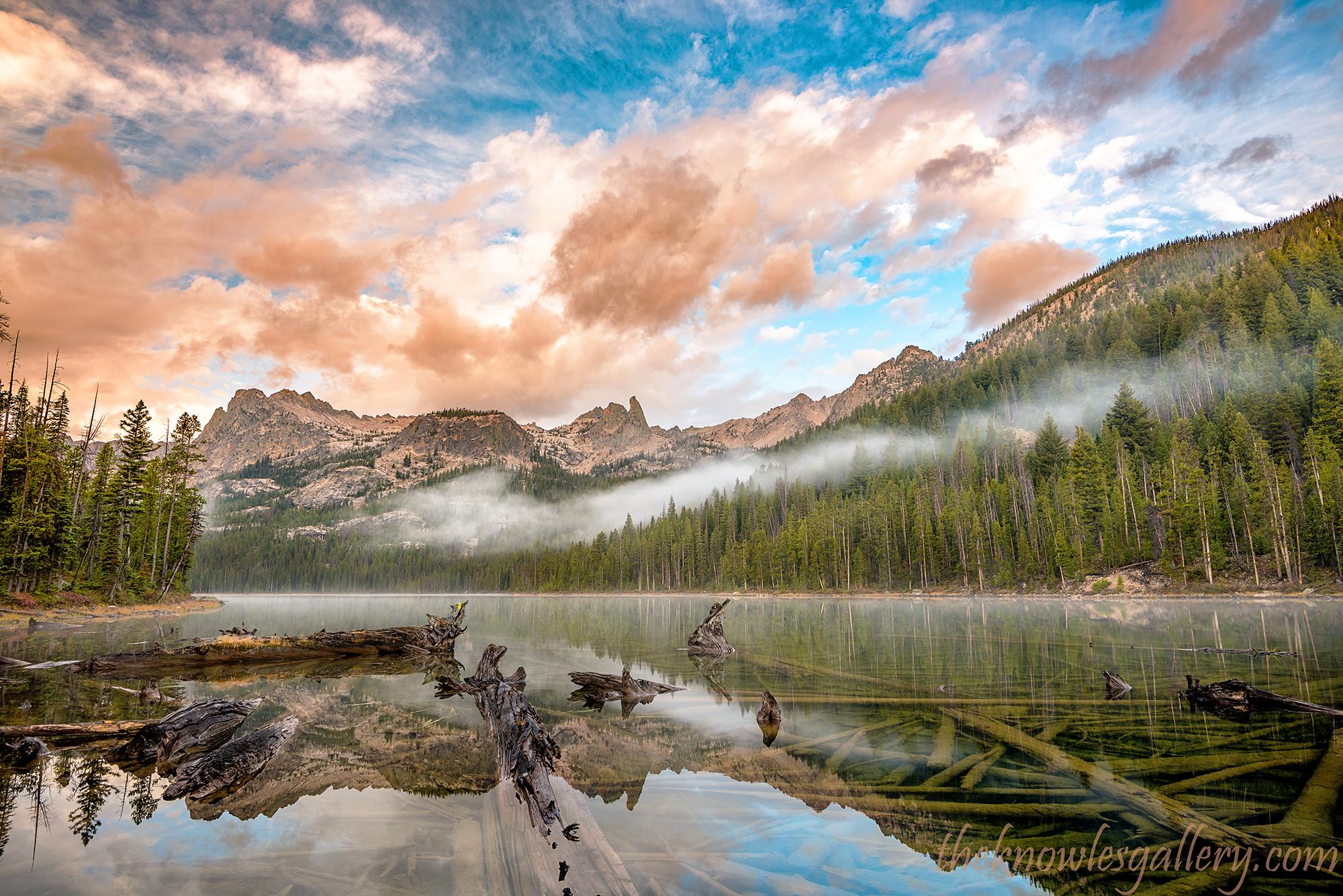 Serene lake reflecting mountains and forest under a colorful, cloudy sky.