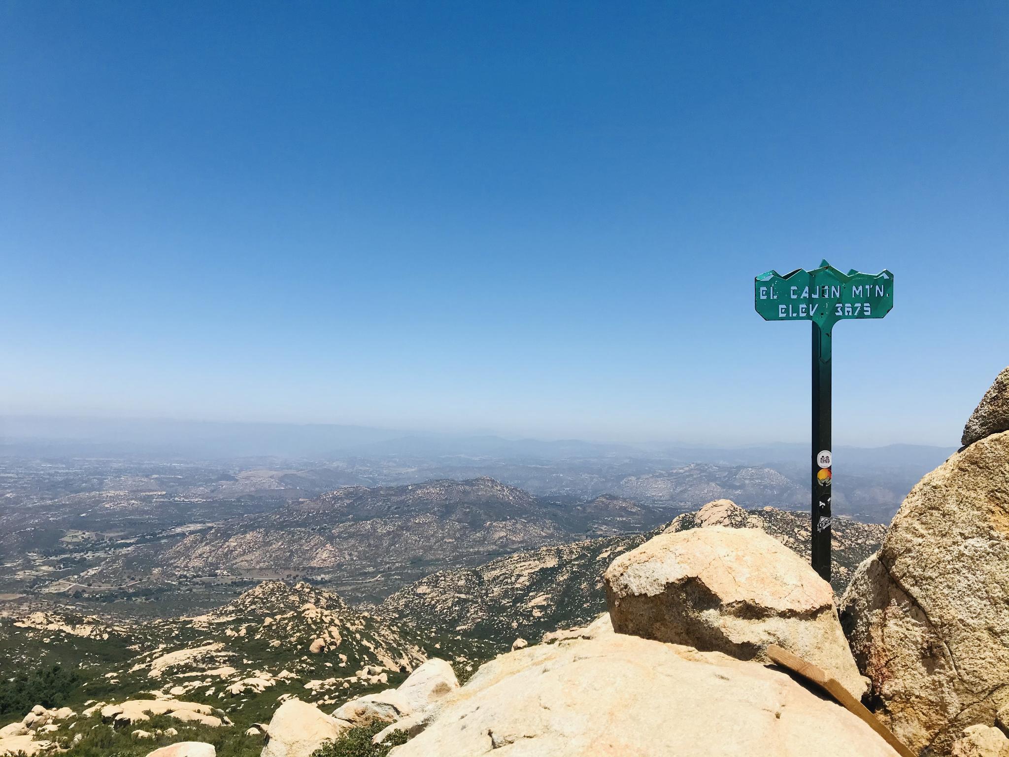 Green sign on a rocky peak overlooking a vast, hazy valley.