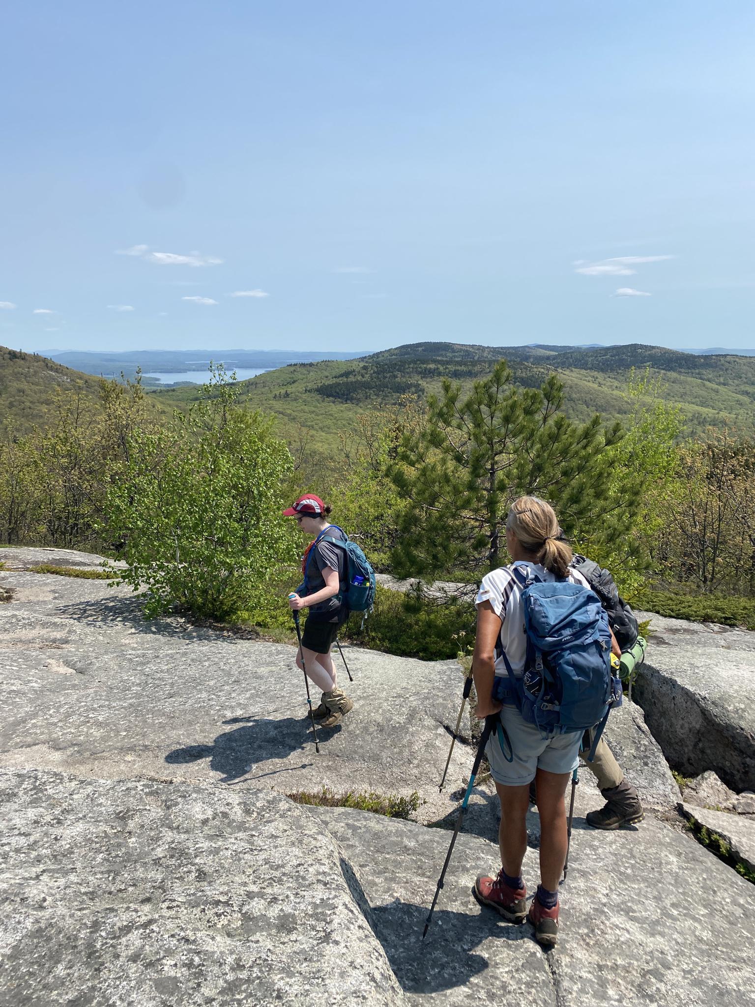 Three hikers with poles on a rocky overlook, enjoying sunny mountain views.