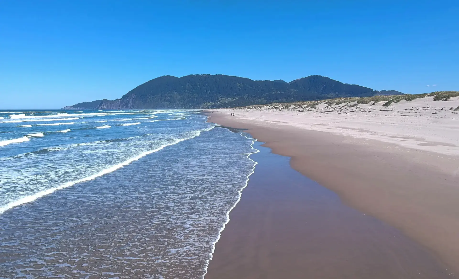 Sunny day on a wide sandy beach with ocean waves and distant green mountains.
