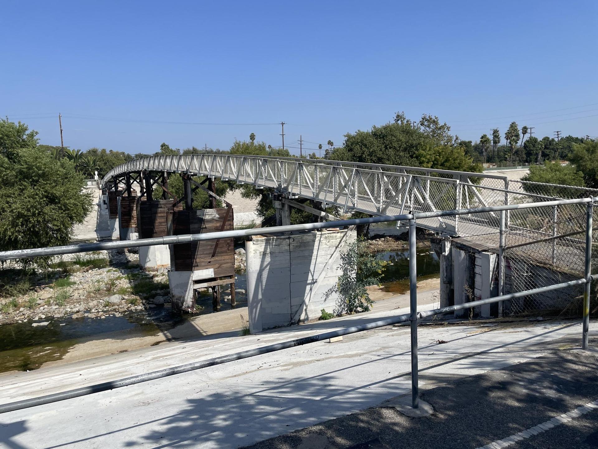 A white pedestrian bridge spans a river with concrete banks, surrounded by trees.