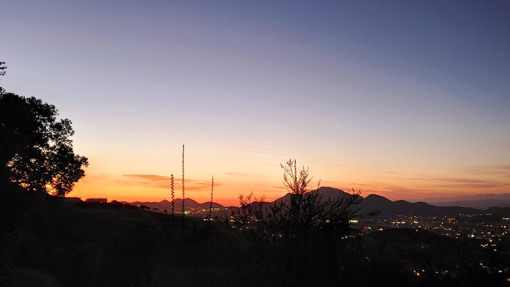 Sunset over distant mountains and a city, silhouetted trees.