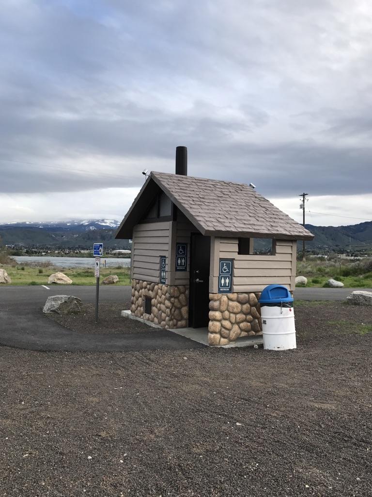 Small rustic restroom building on gravel, with distant snowy mountains under cloudy sky.