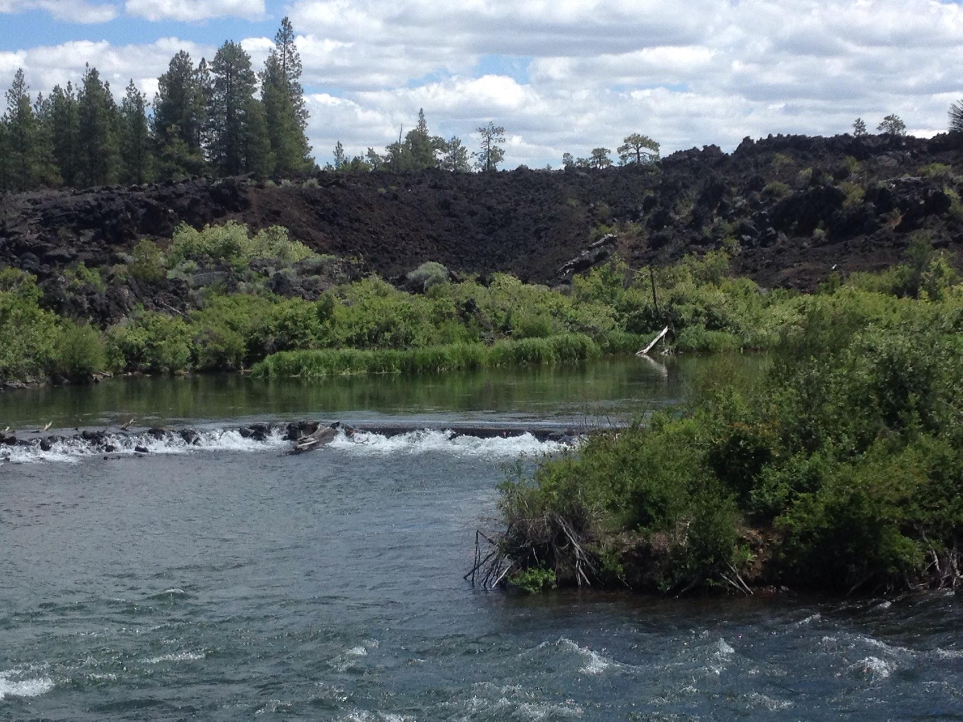 River flowing past green bushes and dark, rocky hills under a cloudy sky.