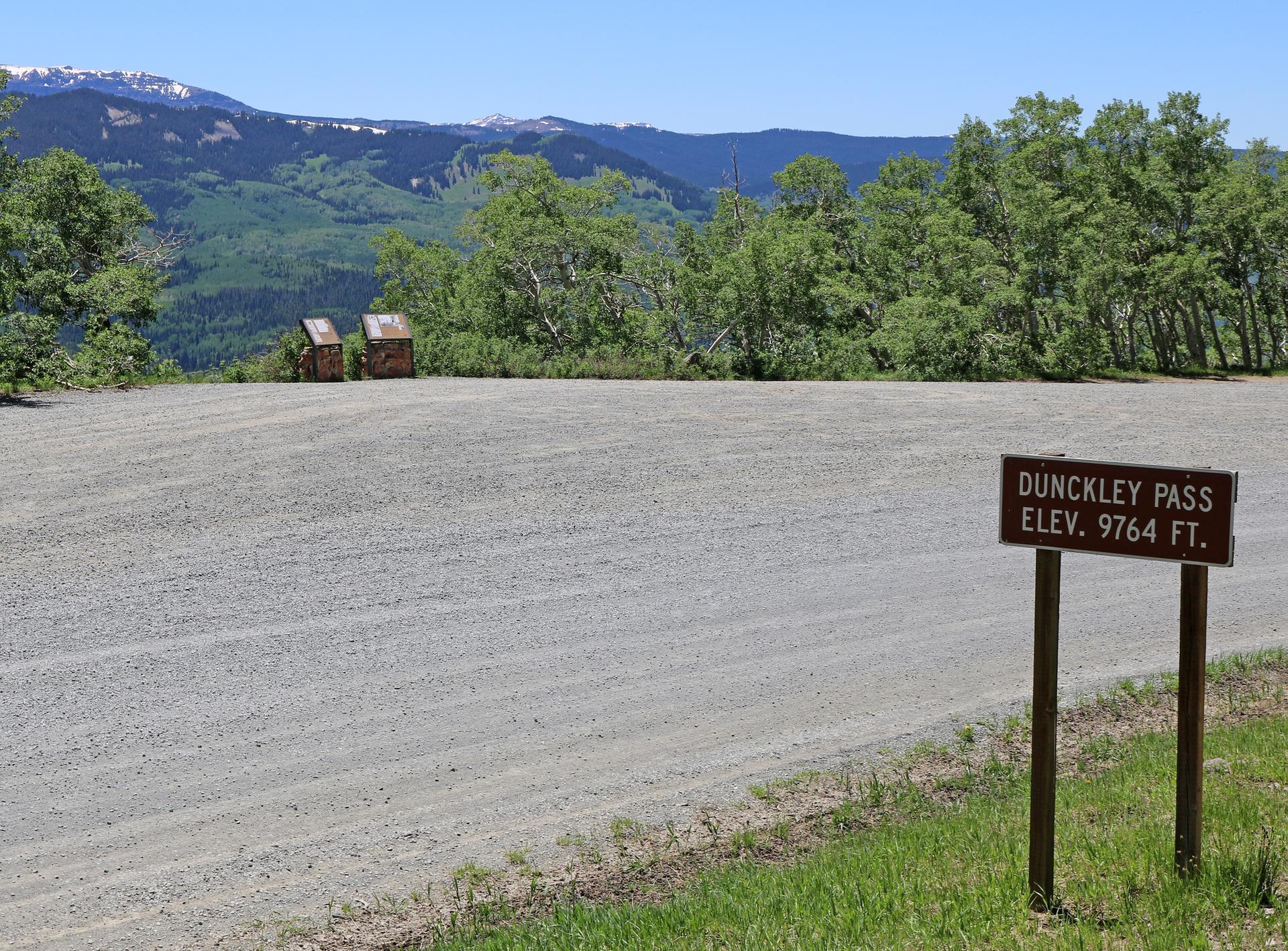 Gravel parking area with a sign, overlooking green mountains under a clear blue sky.