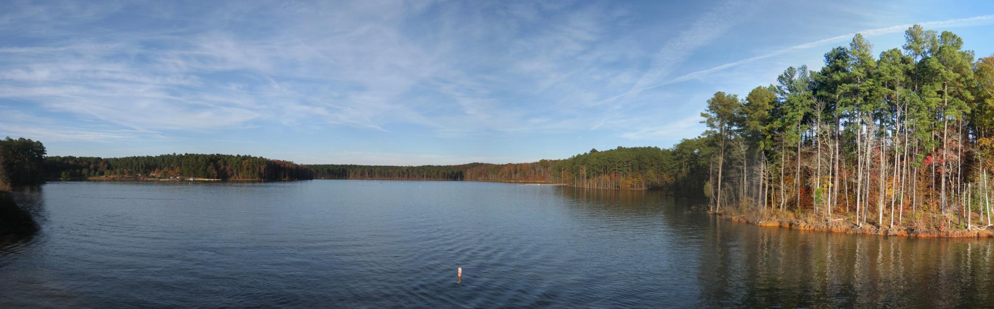 Panoramic view of a calm lake surrounded by dense forests under a bright sky.