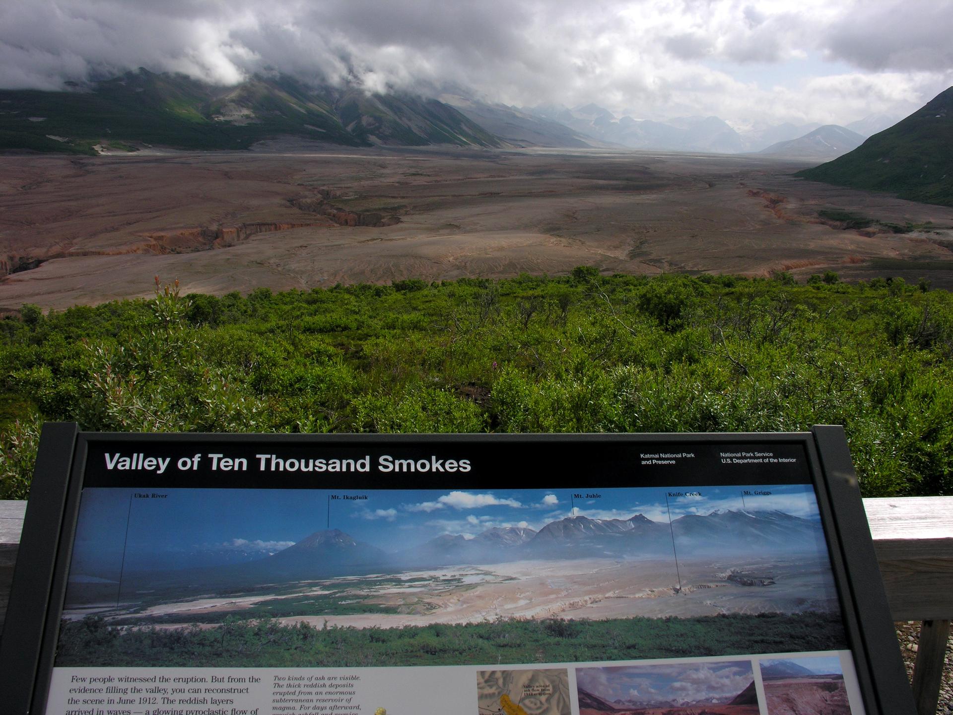 Overcast view of a vast, barren valley with mountains and a descriptive sign.