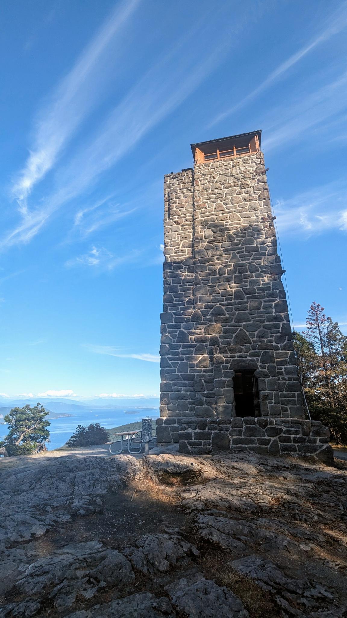 Tall stone tower on a rocky outcrop overlooking distant water under a bright sky.