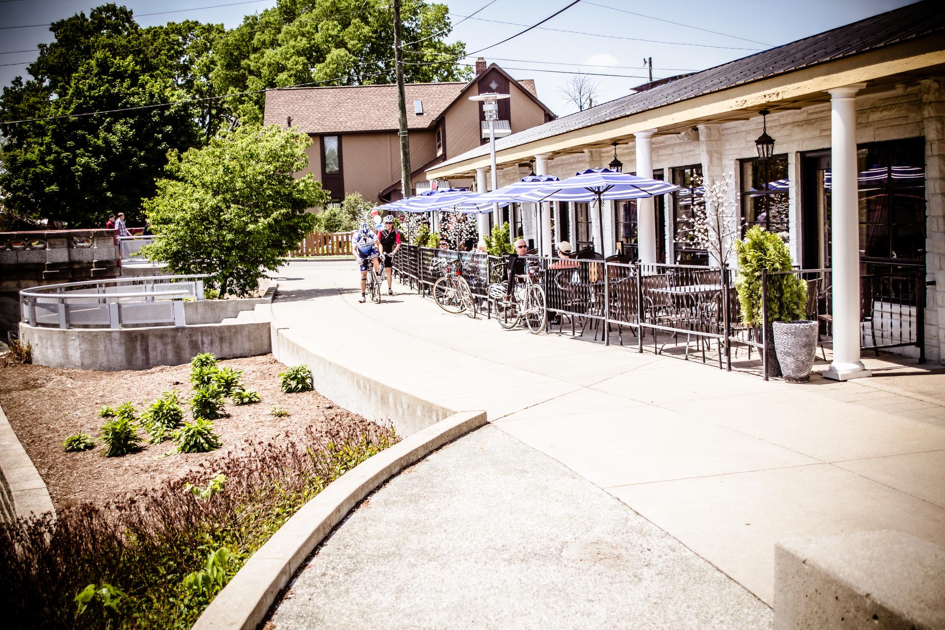 Cyclists ride past an outdoor cafe with blue umbrellas on a bright day.