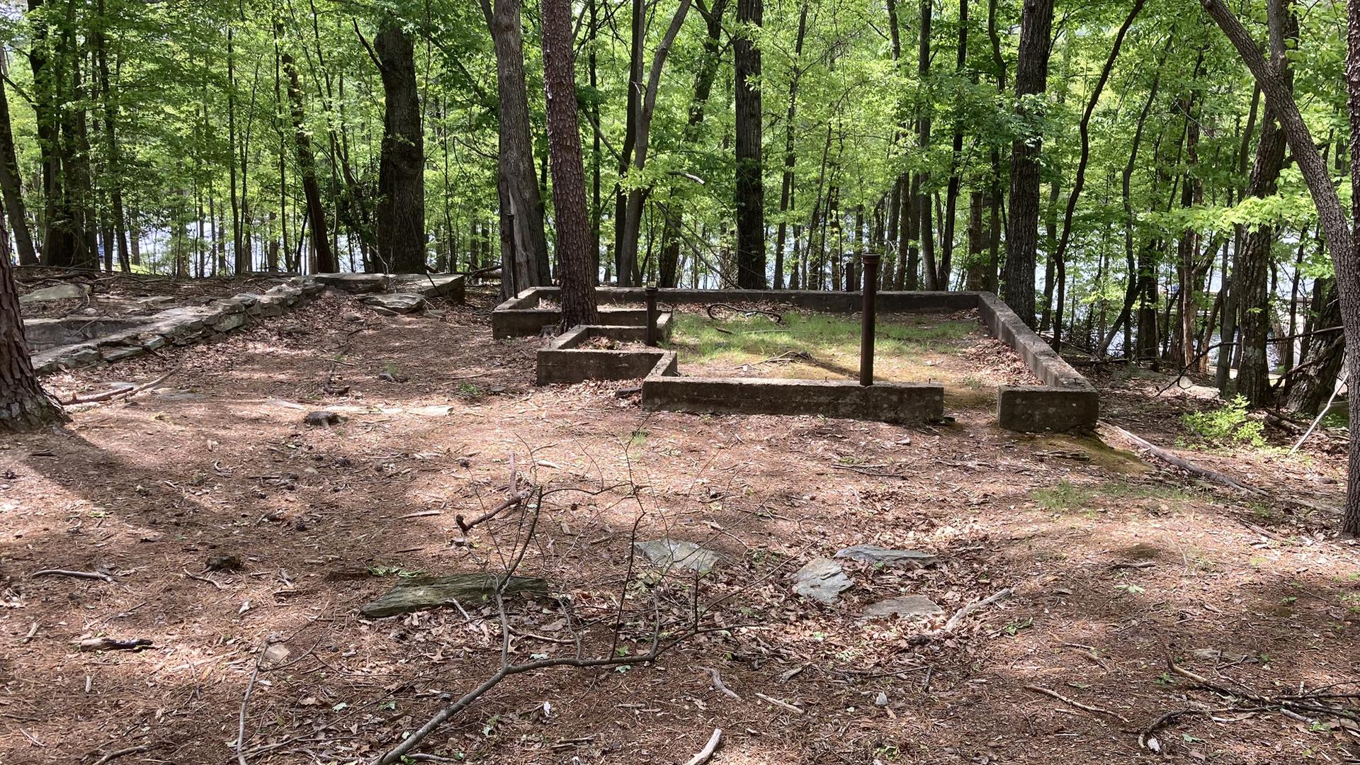 Concrete foundations in a sunny forest with a lake visible through trees.