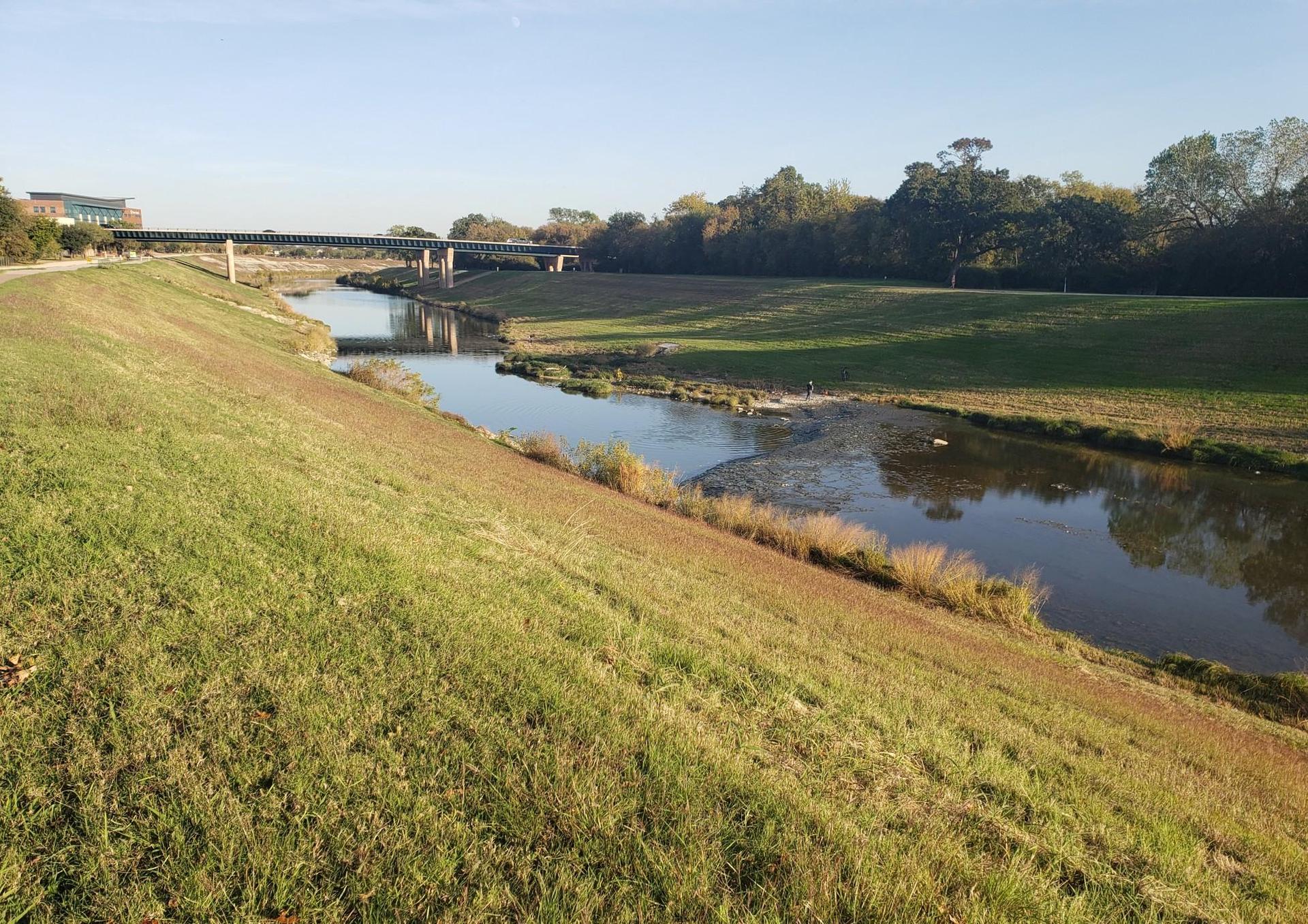 Sunny river flowing between steep grassy banks with a distant bridge.