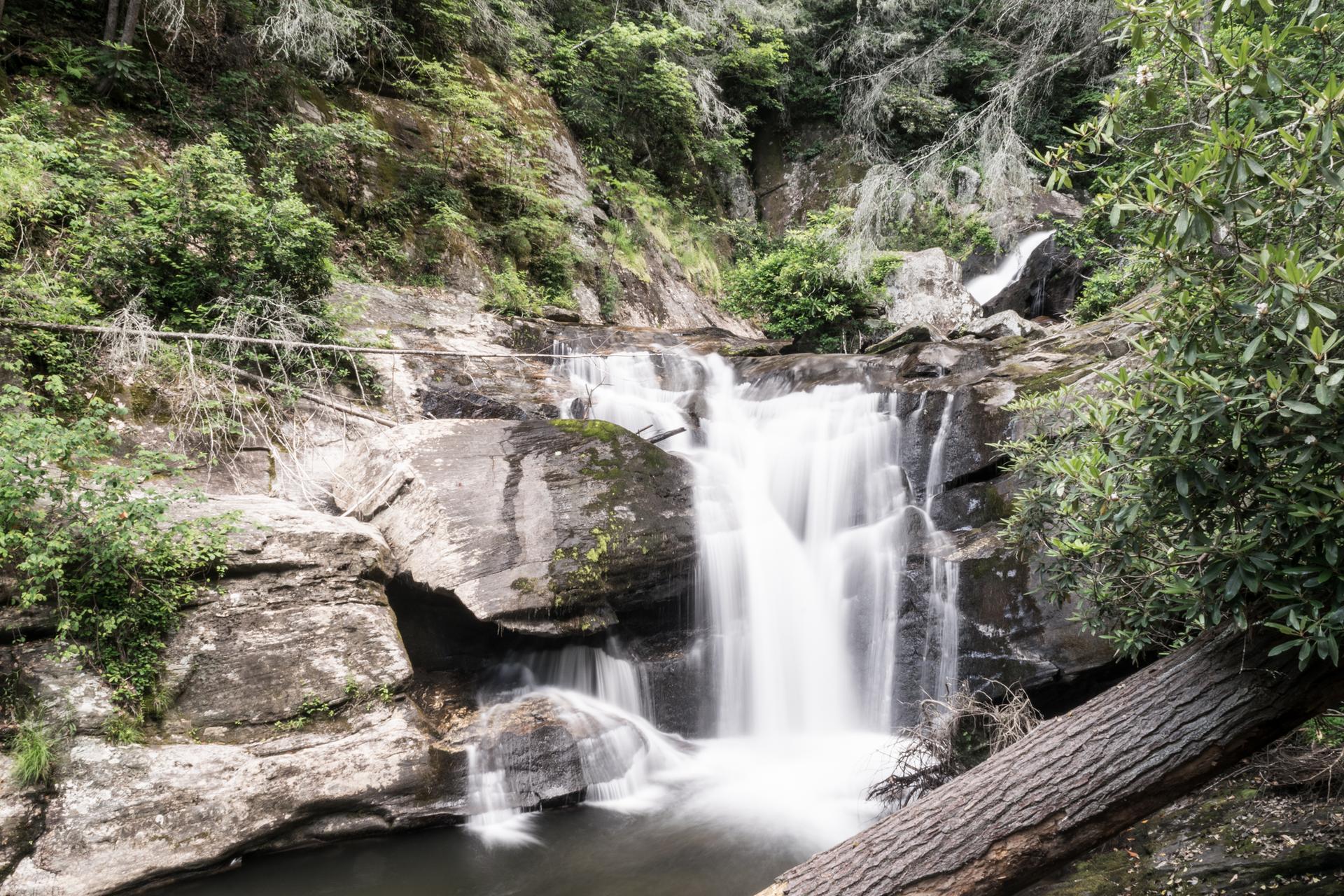 Long exposure waterfall flowing over rocks, surrounded by green trees and a fallen log.