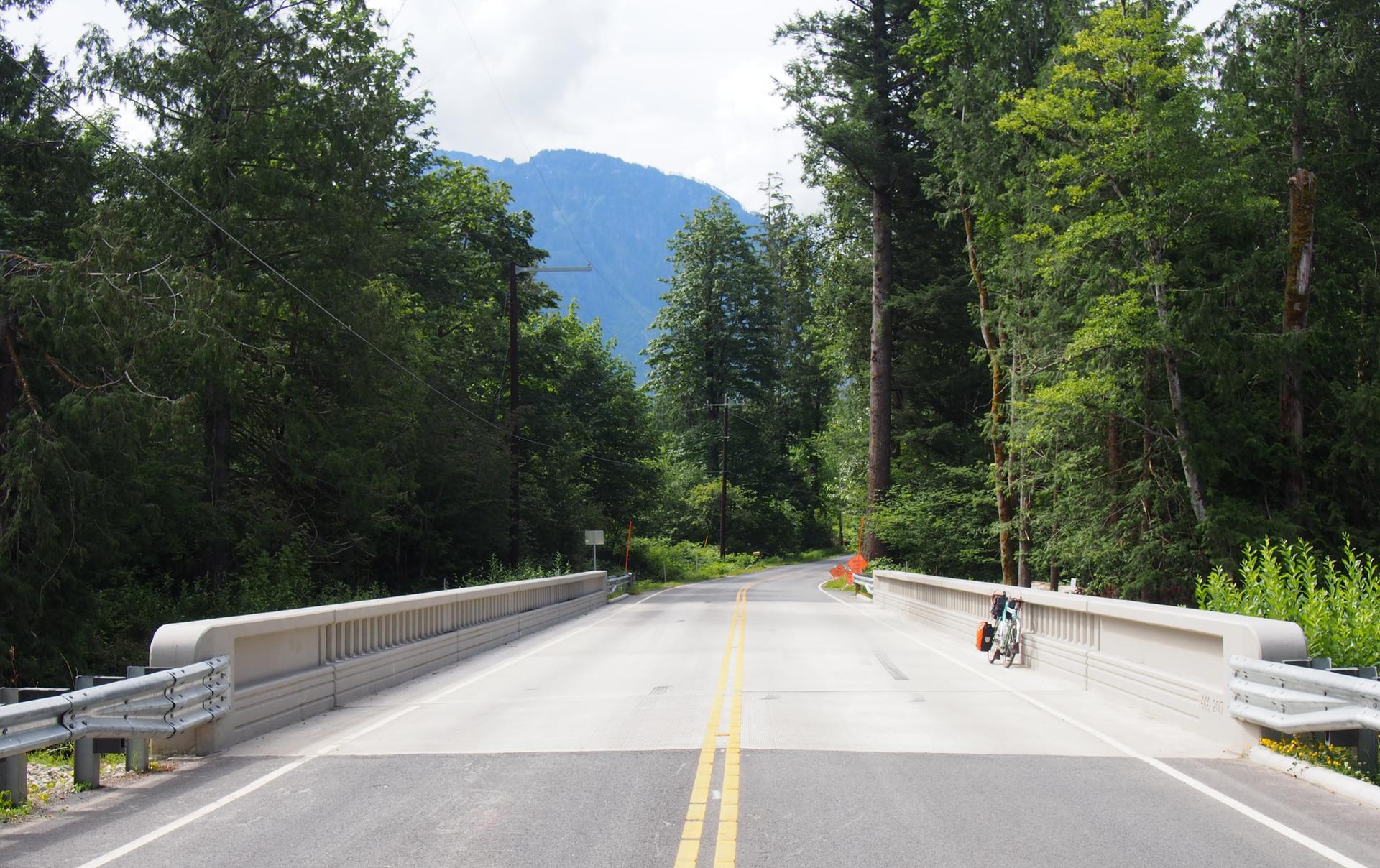 A bicycle parked on a bridge over a road, surrounded by forest and mountains.