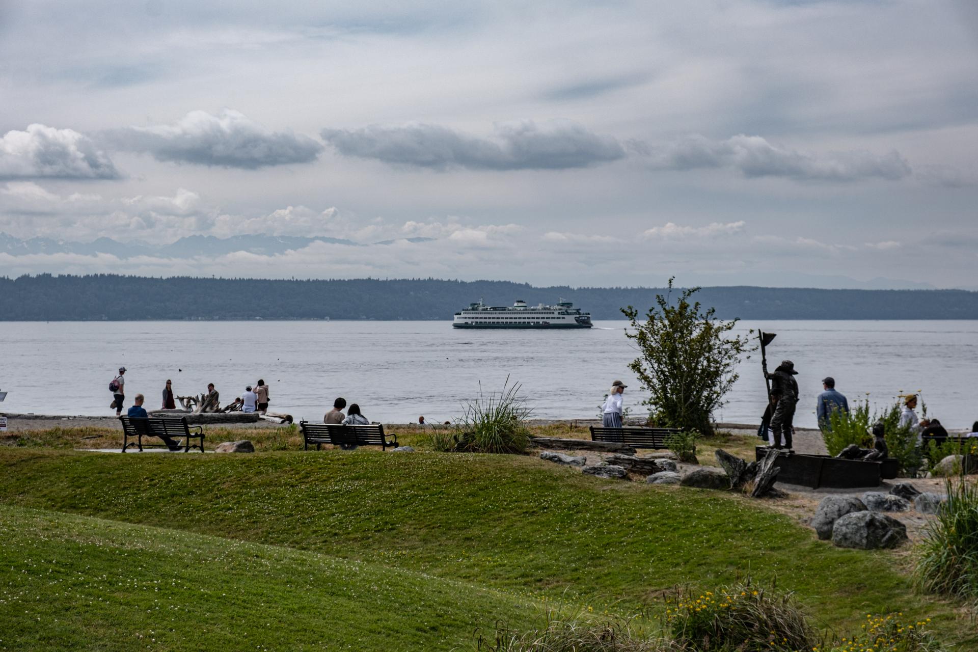 People relax on a grassy bank and beach with a ferry on cloudy water.