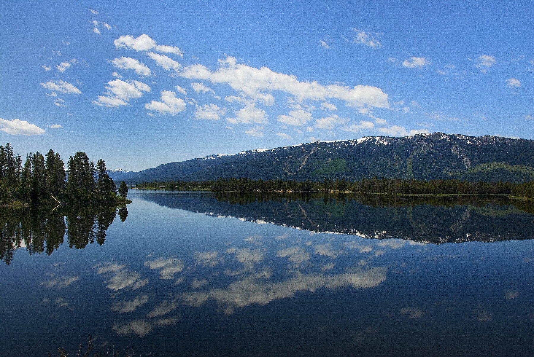 Calm lake reflecting blue sky, white clouds, and distant snow-capped mountains.
