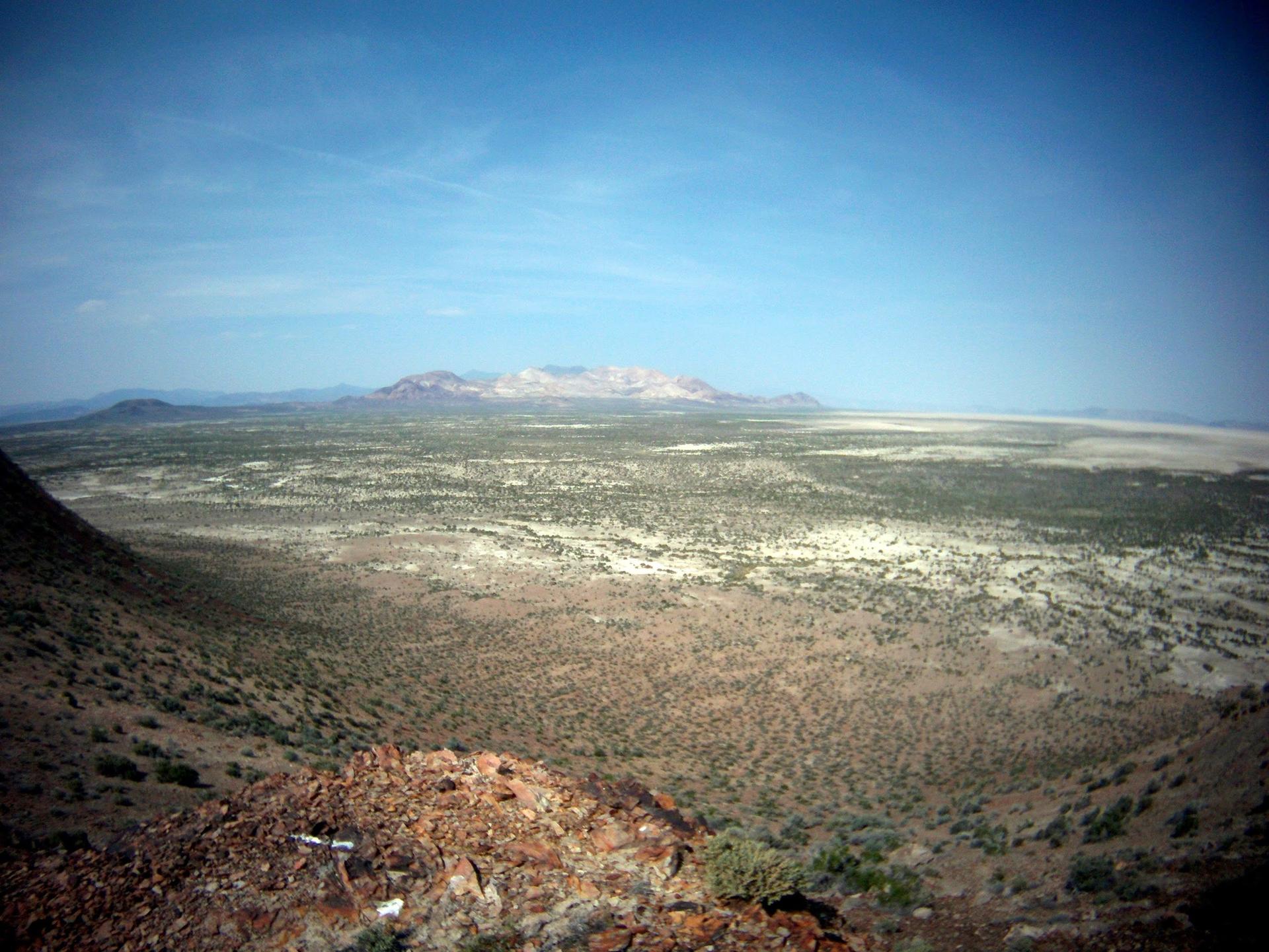Vast desert landscape with scrub brush, distant mountains, and a clear blue sky.