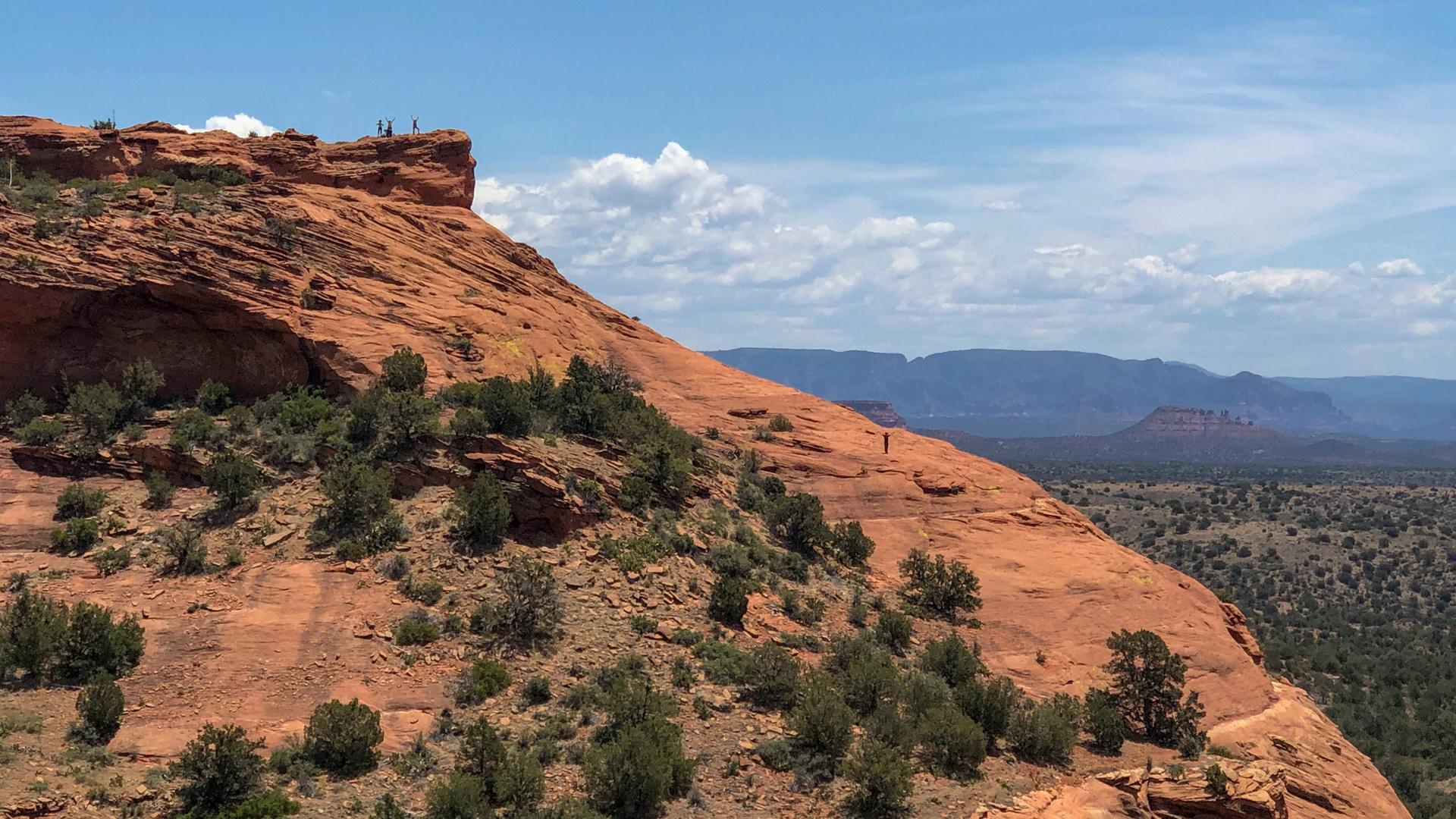 People hiking on a massive red rock formation under a bright sky.