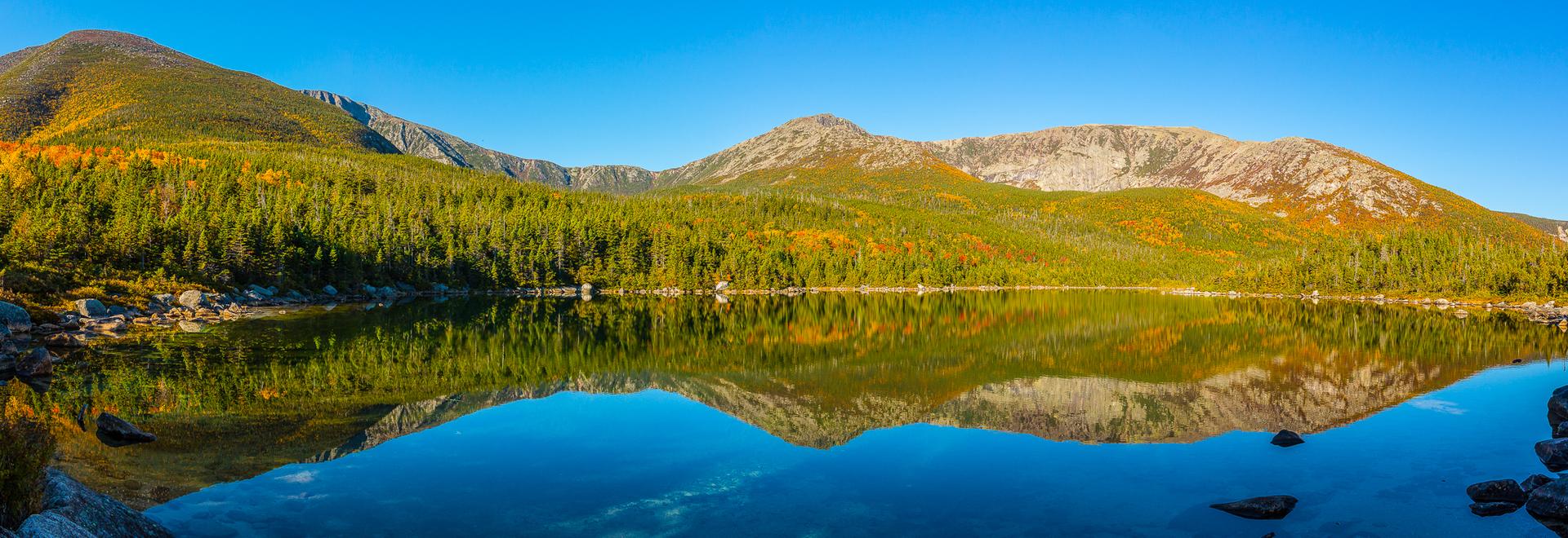 Panoramic view of a calm lake reflecting sunlit mountains and autumn forest.