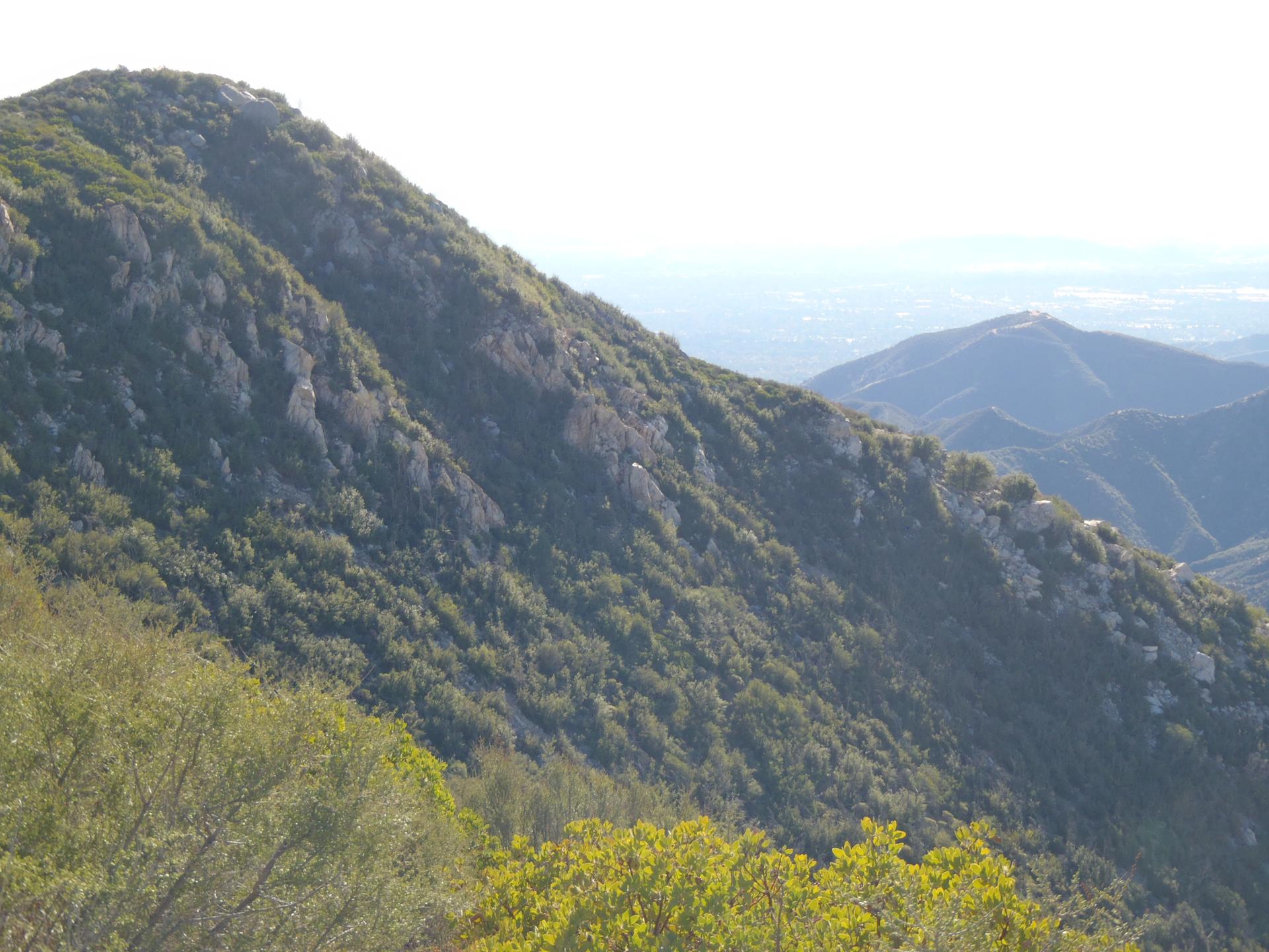 Steep, green mountain slope under bright daylight with distant hazy mountains.