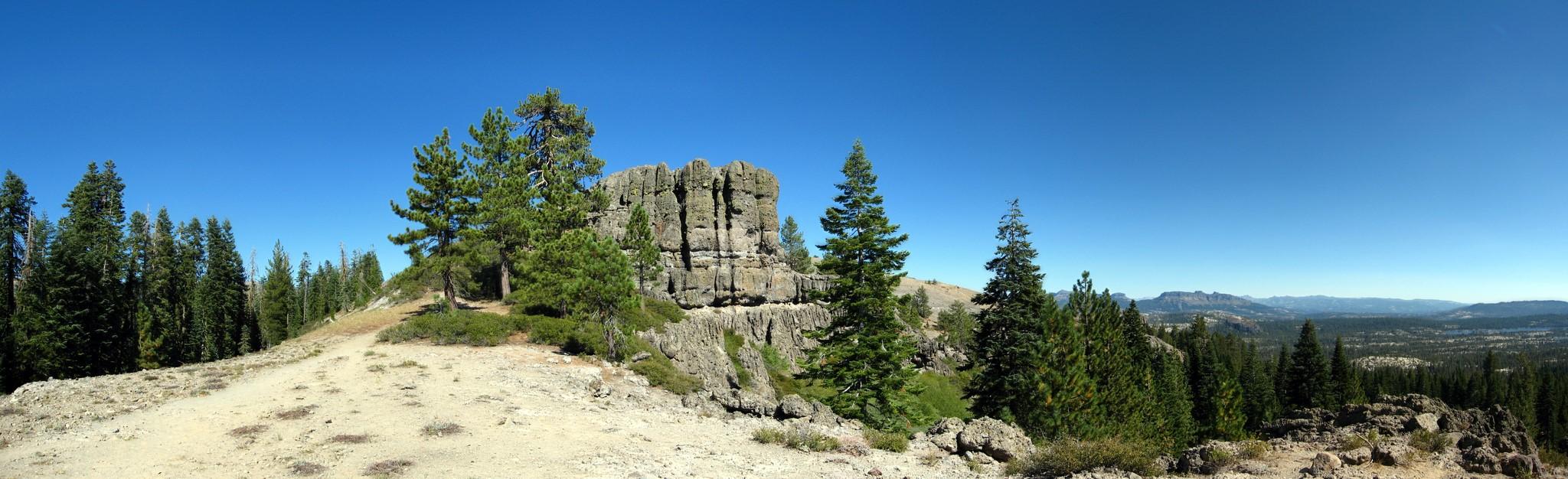 Panoramic view of a rocky outcrop, pine trees, and a dirt path under a clear sky.
