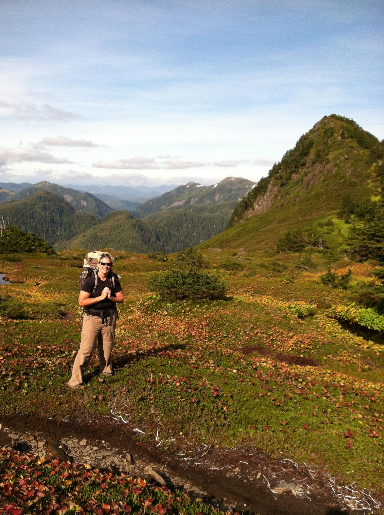 Hiker with backpack standing on a grassy hillside with distant mountains.